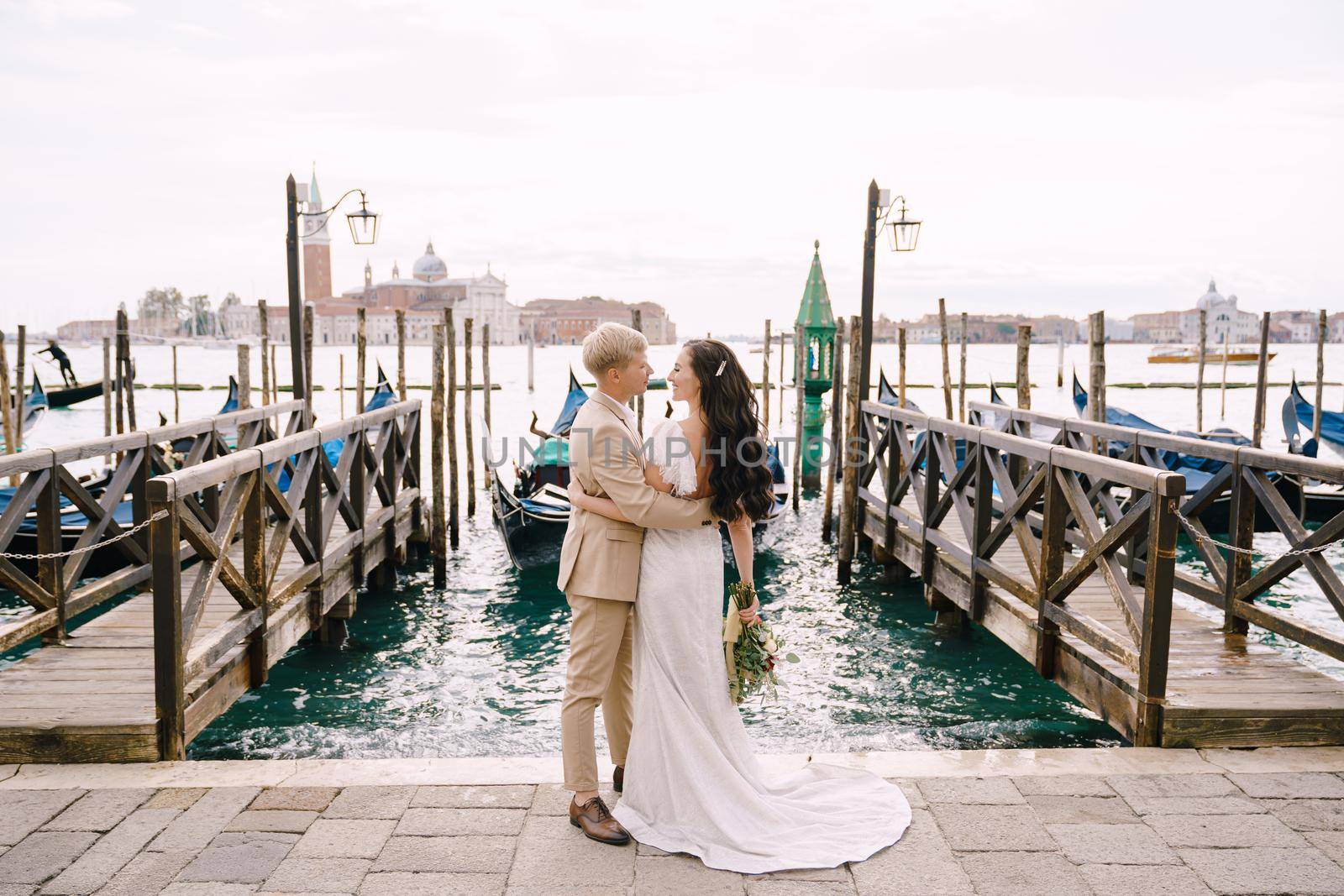 The groom and stand next to the pier for gondolas, hugging, in Venice, near the Piazza San Marco, overlooking San Giorgio Maggiore and the sunset sky. The largest pier for gondolas in Venice, Italy.