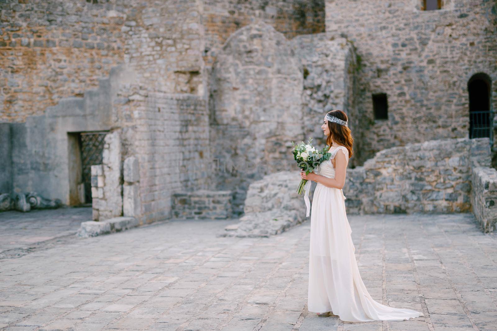 The bride stands on the square near the castle walls in the old town of Budva and holds a bouquet in her hands by Nadtochiy