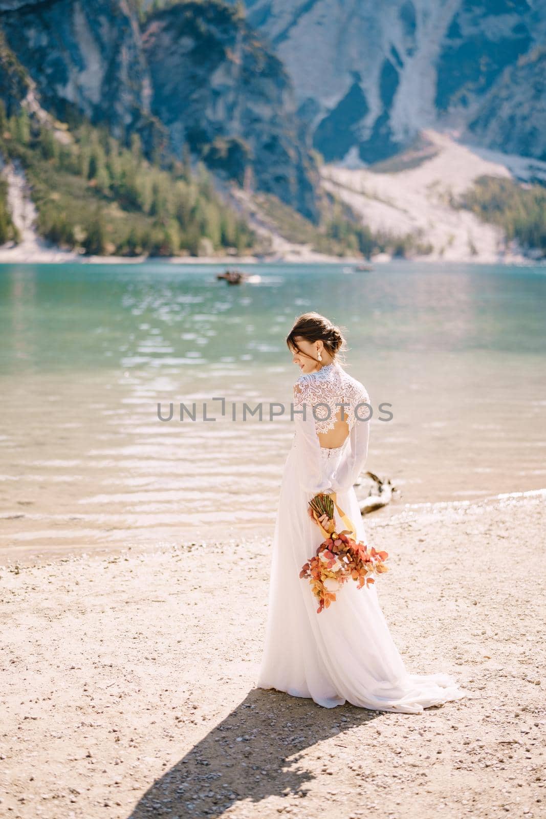Beautiful bride in a white dress with sleeves and lace, with a yellow autumn bouquet of dried flowers and peony roses, at Lago di Braies in Italy. Destination wedding in Europe, at Braies lake.