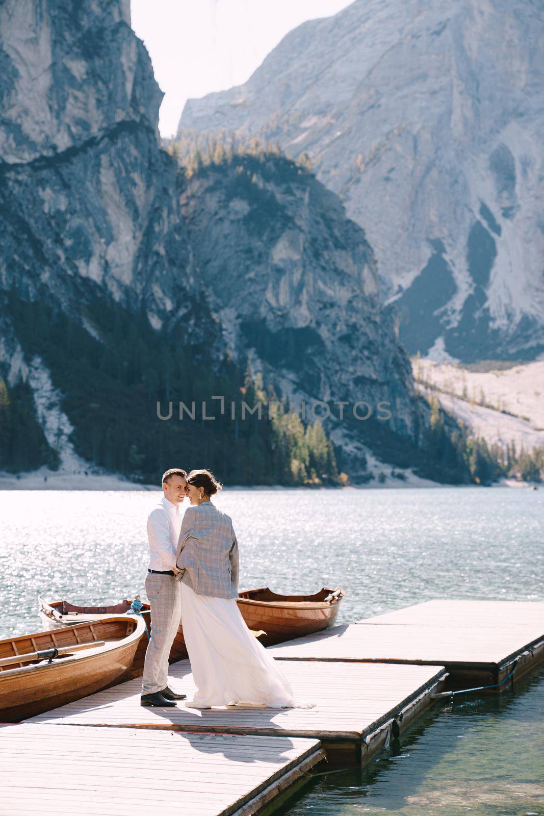 The bride and groom walk along a wooden boat dock at the Lago di Braies in Italy. Wedding in Europe, on Braies lake. Newlyweds walk, kiss, hug on a background of rocky mountains. by Nadtochiy