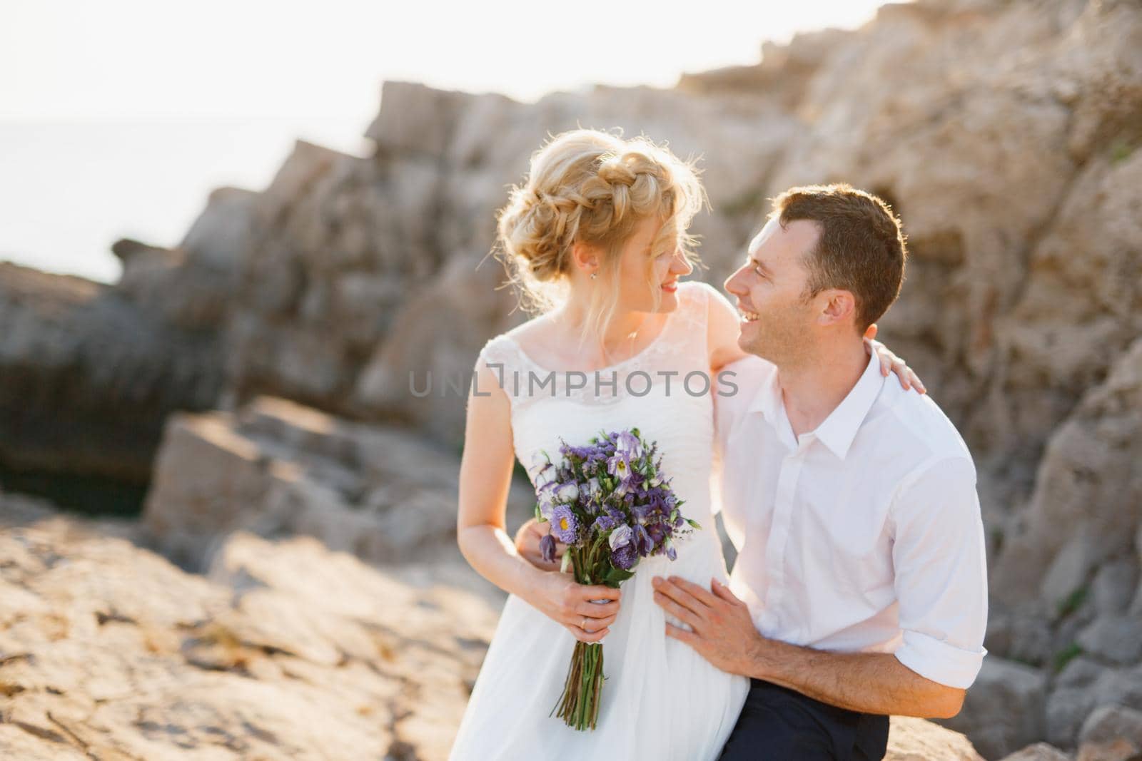 Bride with a bouquet of blue flowers and the groom hug at the rocks by the sea by Nadtochiy