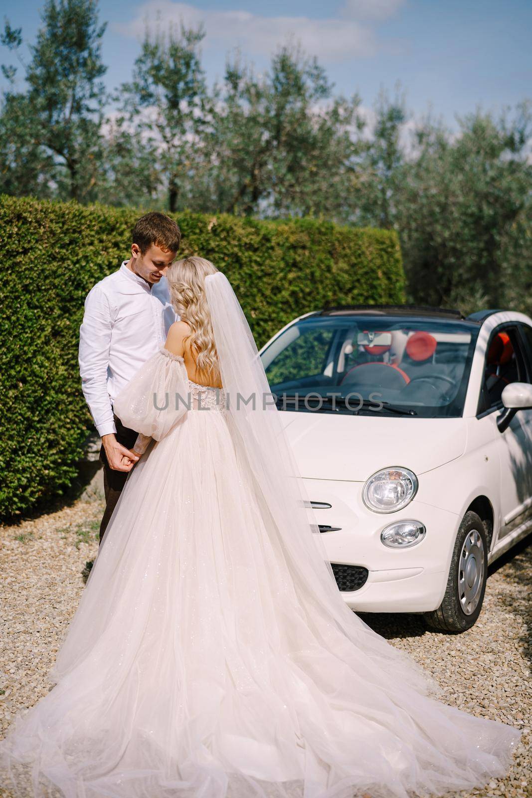 Beautiful bride and groom leaning forehead to forehrad and holding hands in front of a convertible at the old villa in Italy, in Tuscany, near Florence