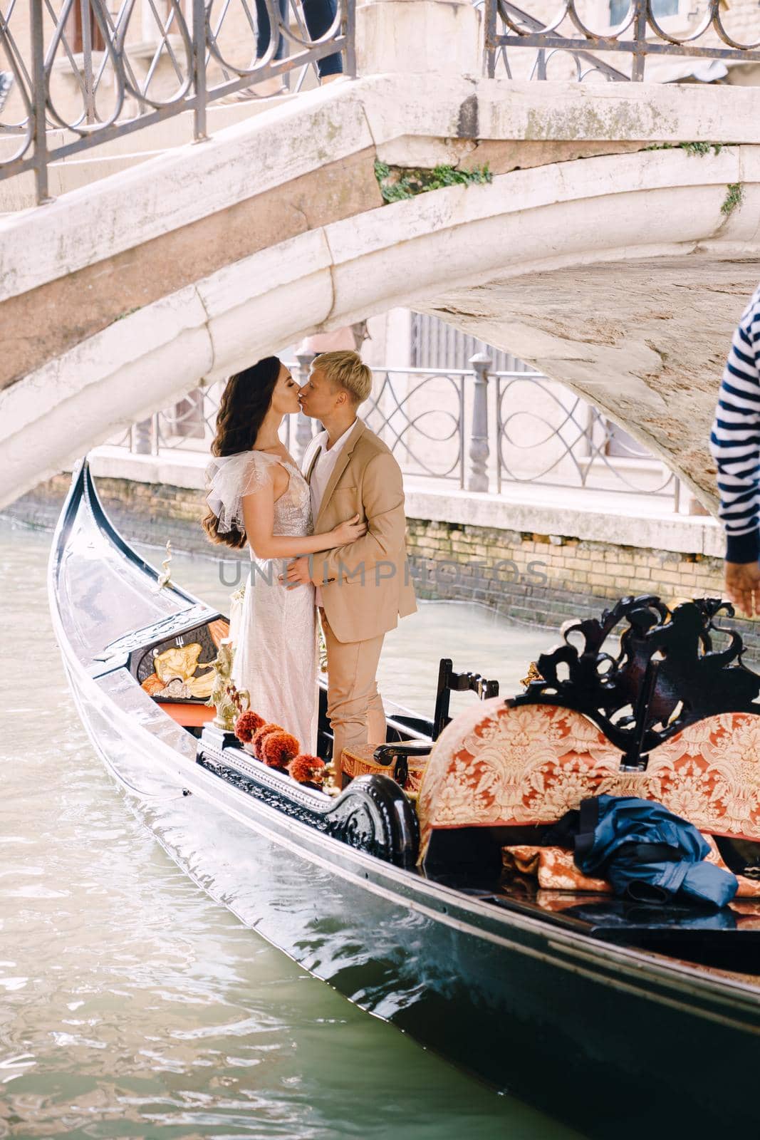 The gondolier rides the bride and groom in a classic wooden gondola along a narrow Venetian canal. The gondola floats under a stone bridge, the newlyweds kiss.