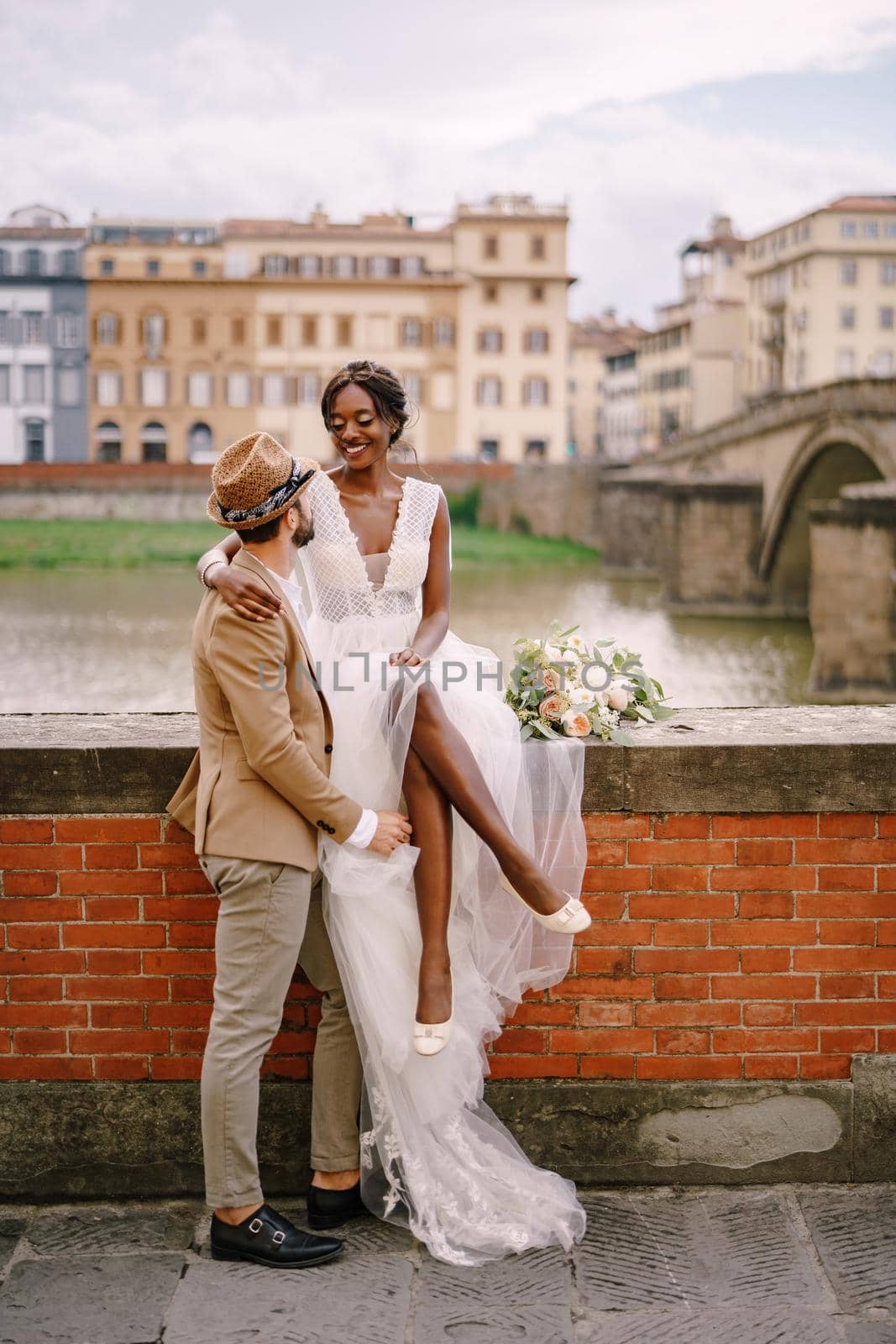 Interracial wedding couple. Wedding in Florence, Italy. An African-American bride is sitting on a brick wall and Caucasian groom is hugging her. Arno River Embankment, overlooking city and bridges by Nadtochiy