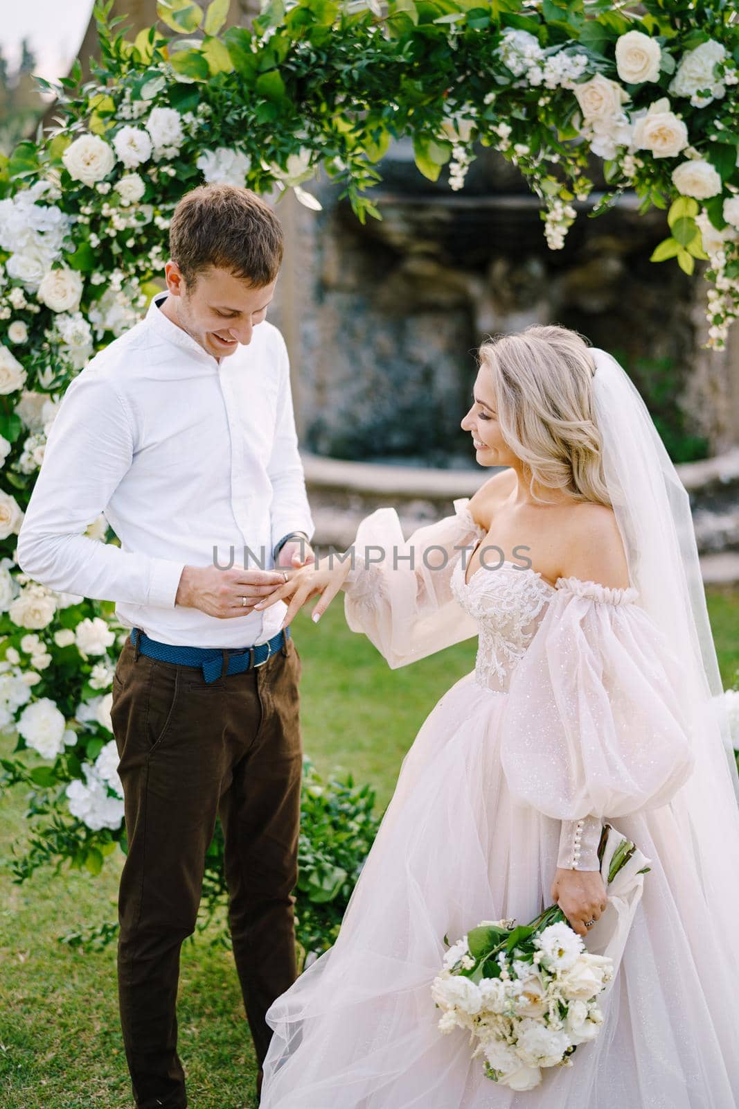 Wedding at an old winery villa in Tuscany, Italy. Round wedding arch decorated with white flowers and greenery in front of an ancient Italian architecture. The groom puts ring on the brides finger.