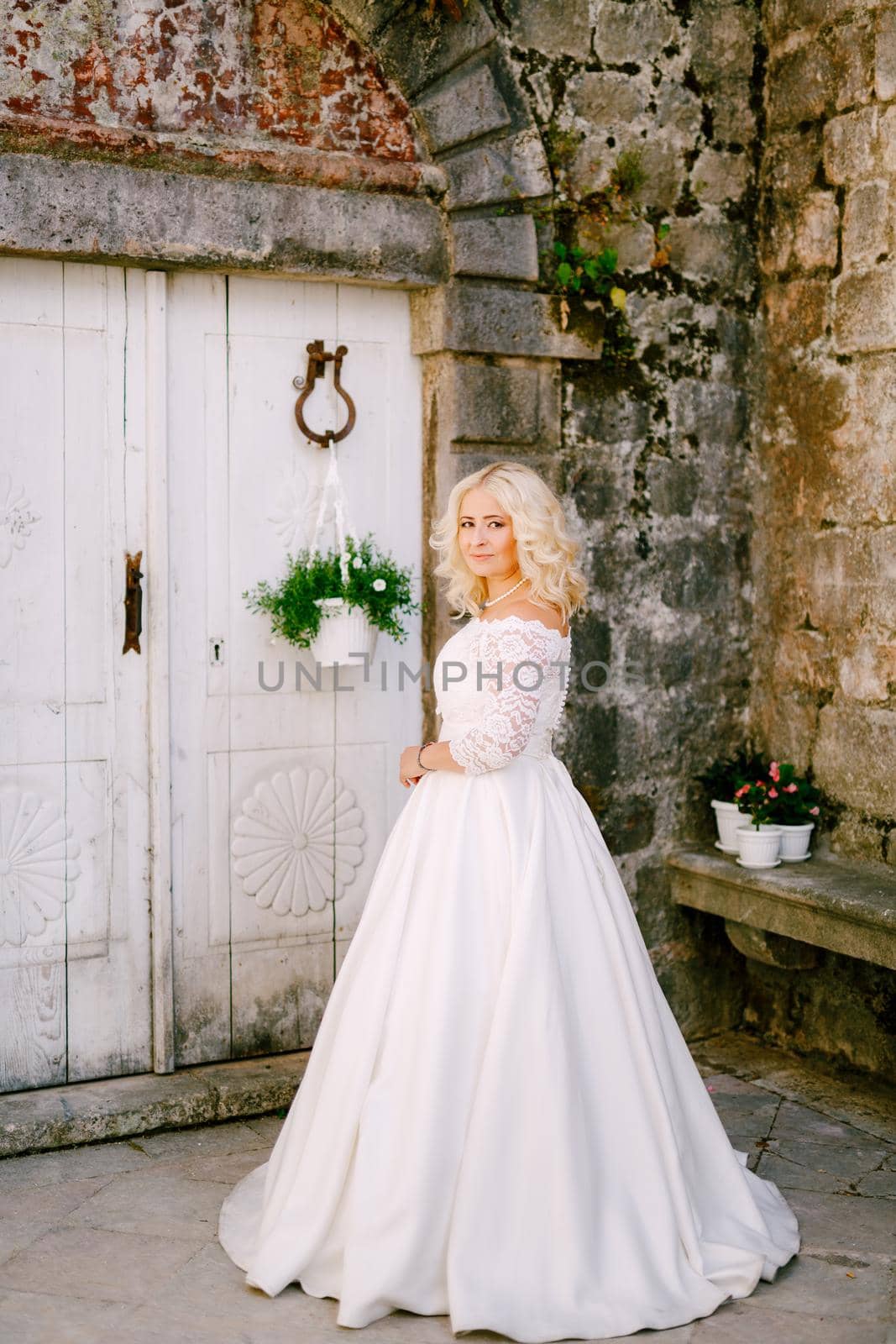 A bride stands in front of an old brick building near a white wooden door with flower pots in Perast by Nadtochiy