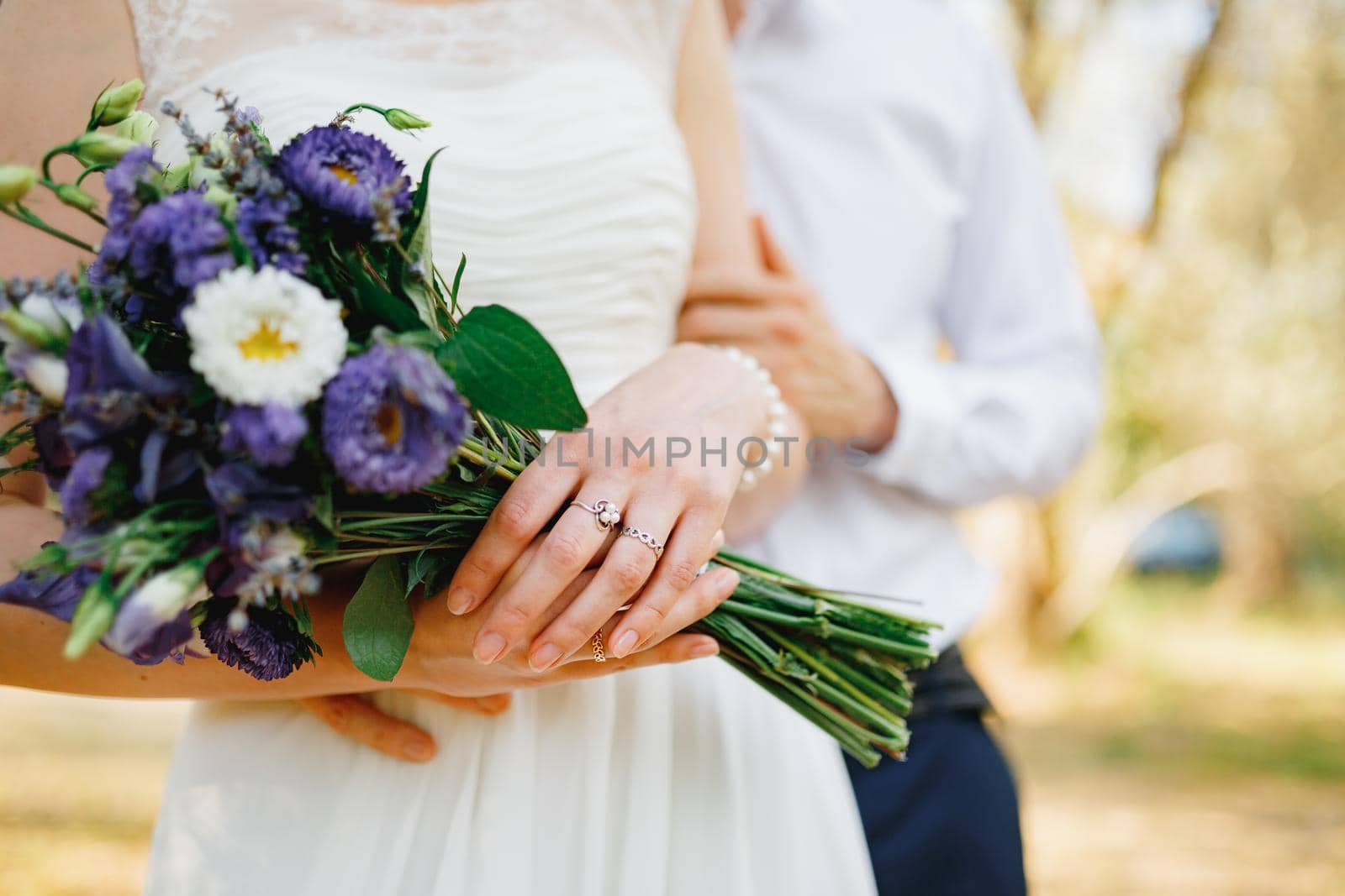 The groom gently hugs the bride in the olive grove, the bride holds a bouquet of blue flowers, close-up. High quality photo