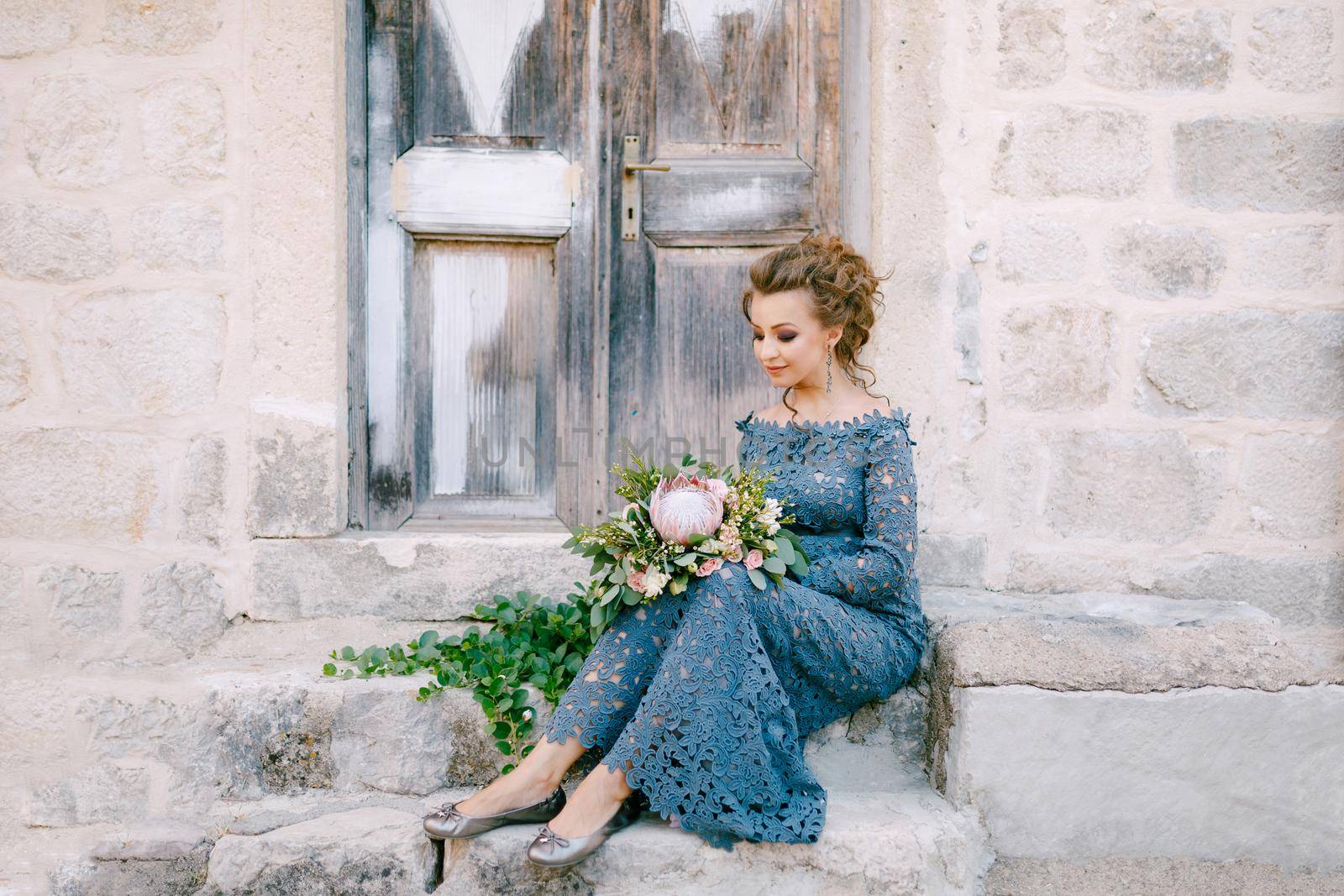 A bride in a stylish blue dress with a bouquet in her hands sits on the steps near an old wooden door by Nadtochiy