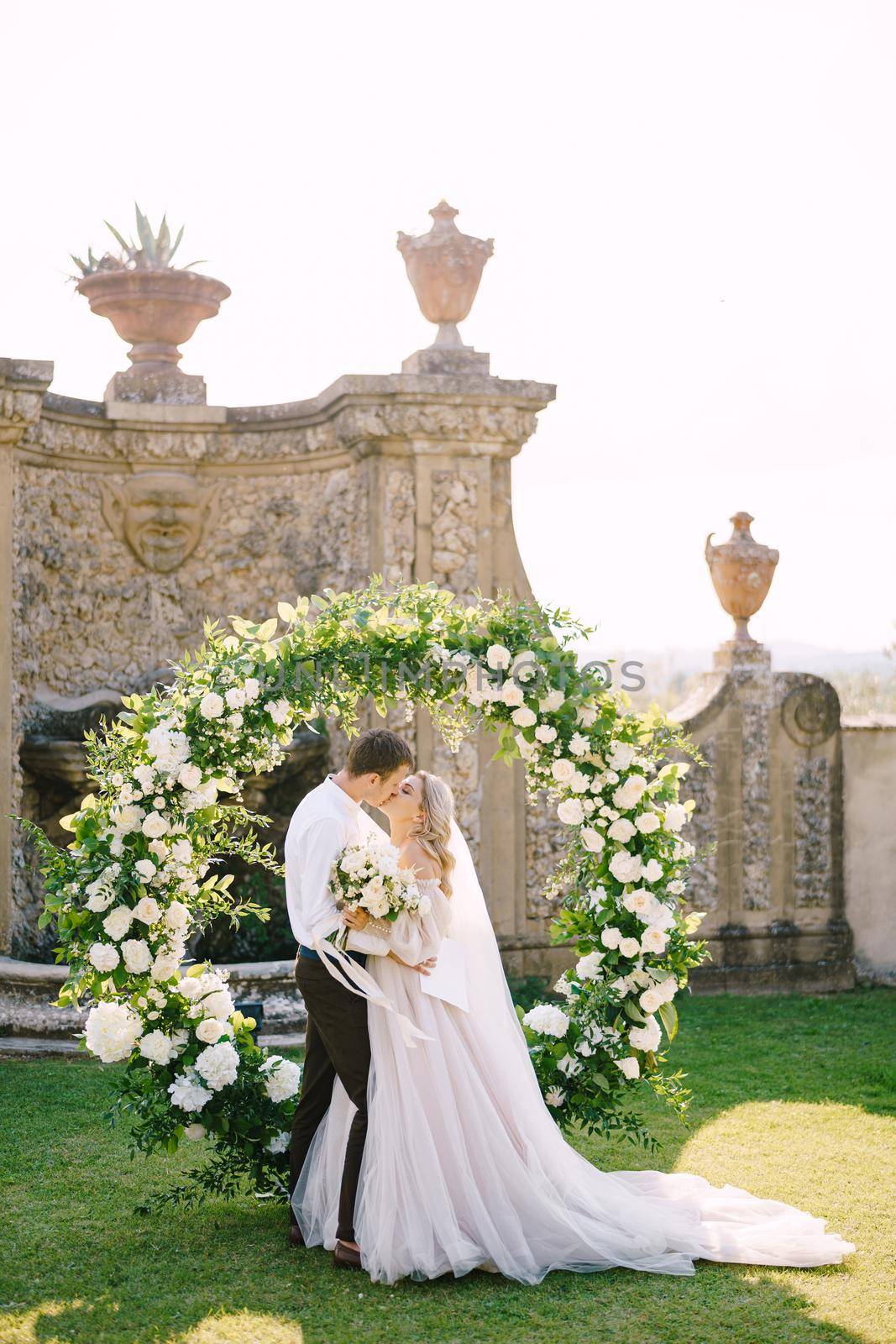 Wedding couple kisses. Wedding at an old winery villa in Tuscany, Italy. Round wedding arch decorated with white flowers and greenery in front of an ancient Italian architecture. by Nadtochiy