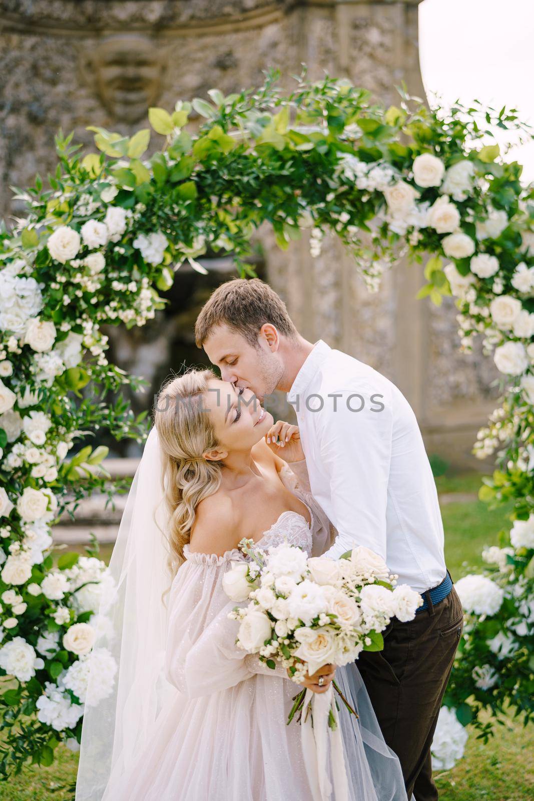 Wedding at an old winery villa in Tuscany, Italy. Round wedding arch decorated with white flowers and greenery in front of an ancient Italian architecture. The wedding couple is kissing.