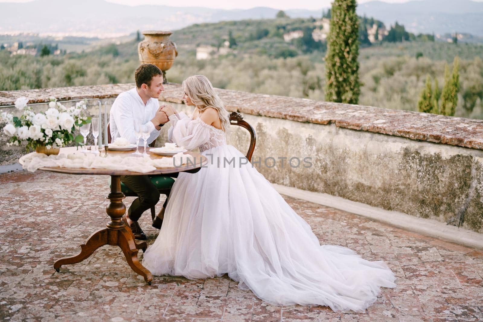 The wedding couple sits at the dinner table on the roof of the old villa, the groom holds the bride's hands. Wedding at an old winery villa in Tuscany, Italy