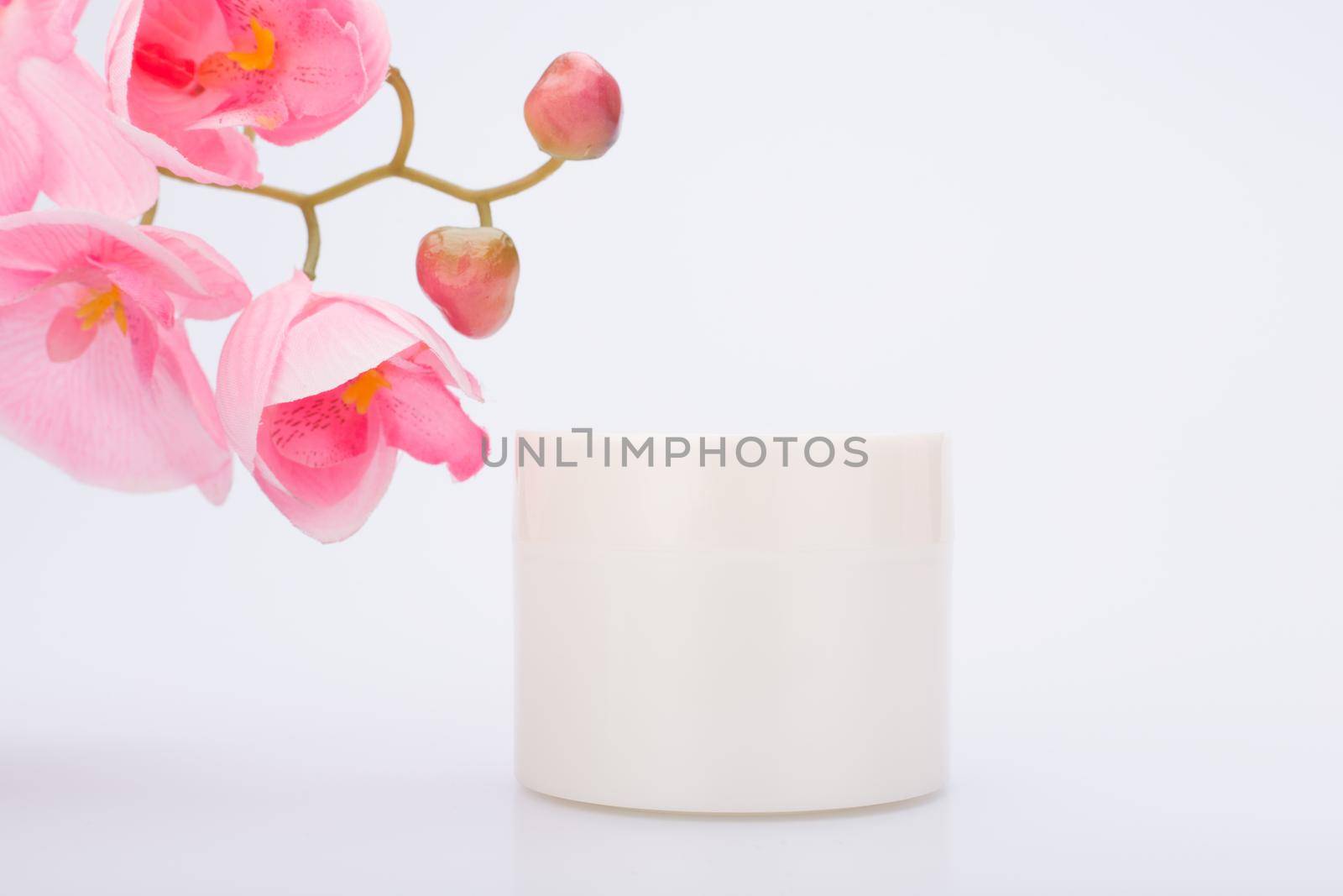 White glossy cream jar against white background with pink flower. Concept of beauty products. Face mask, cream or scrub or hair mask for skin or hair care