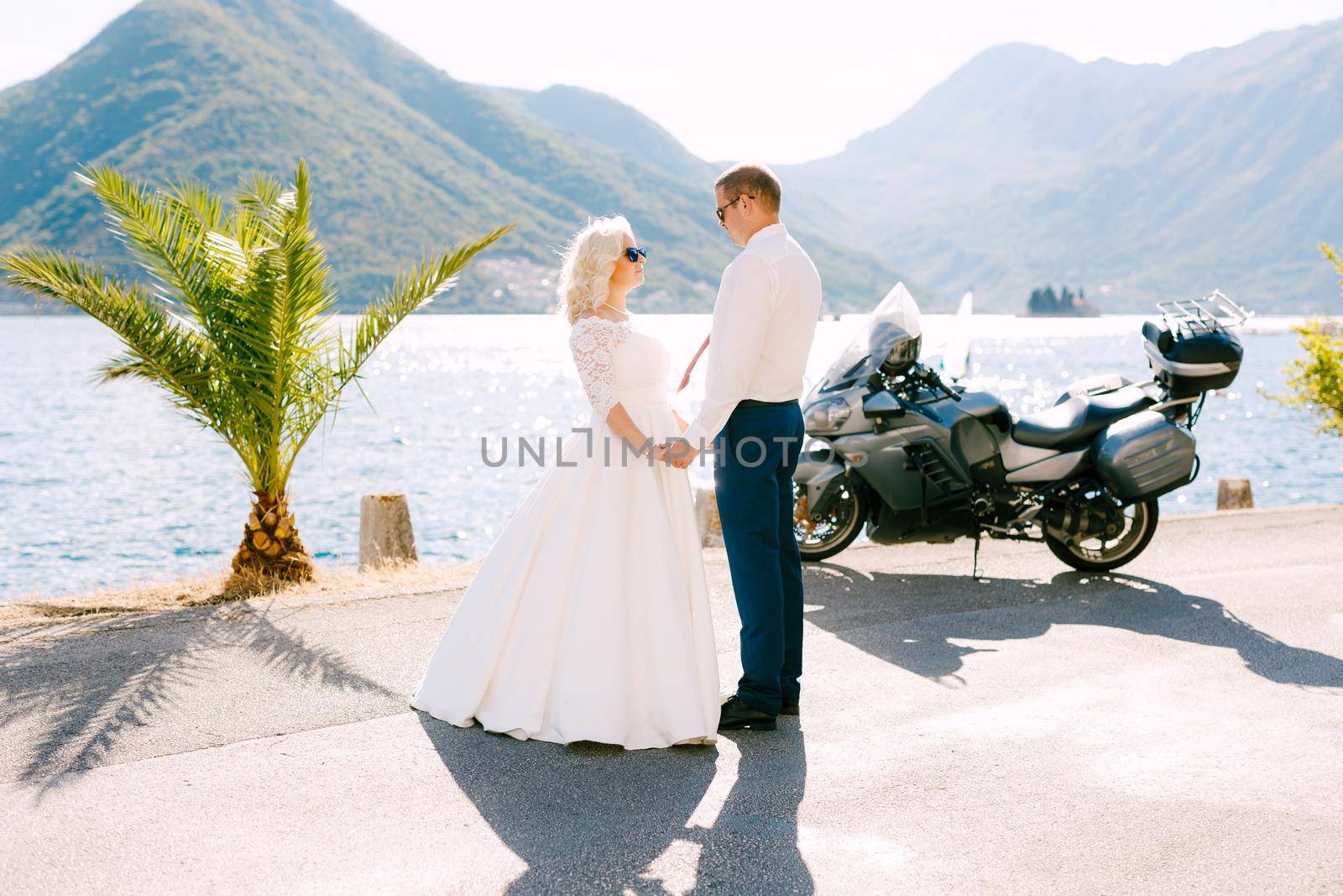 The bride and groom stand on the pier near Perast holding hands, next to them a motorcycle . High quality photo