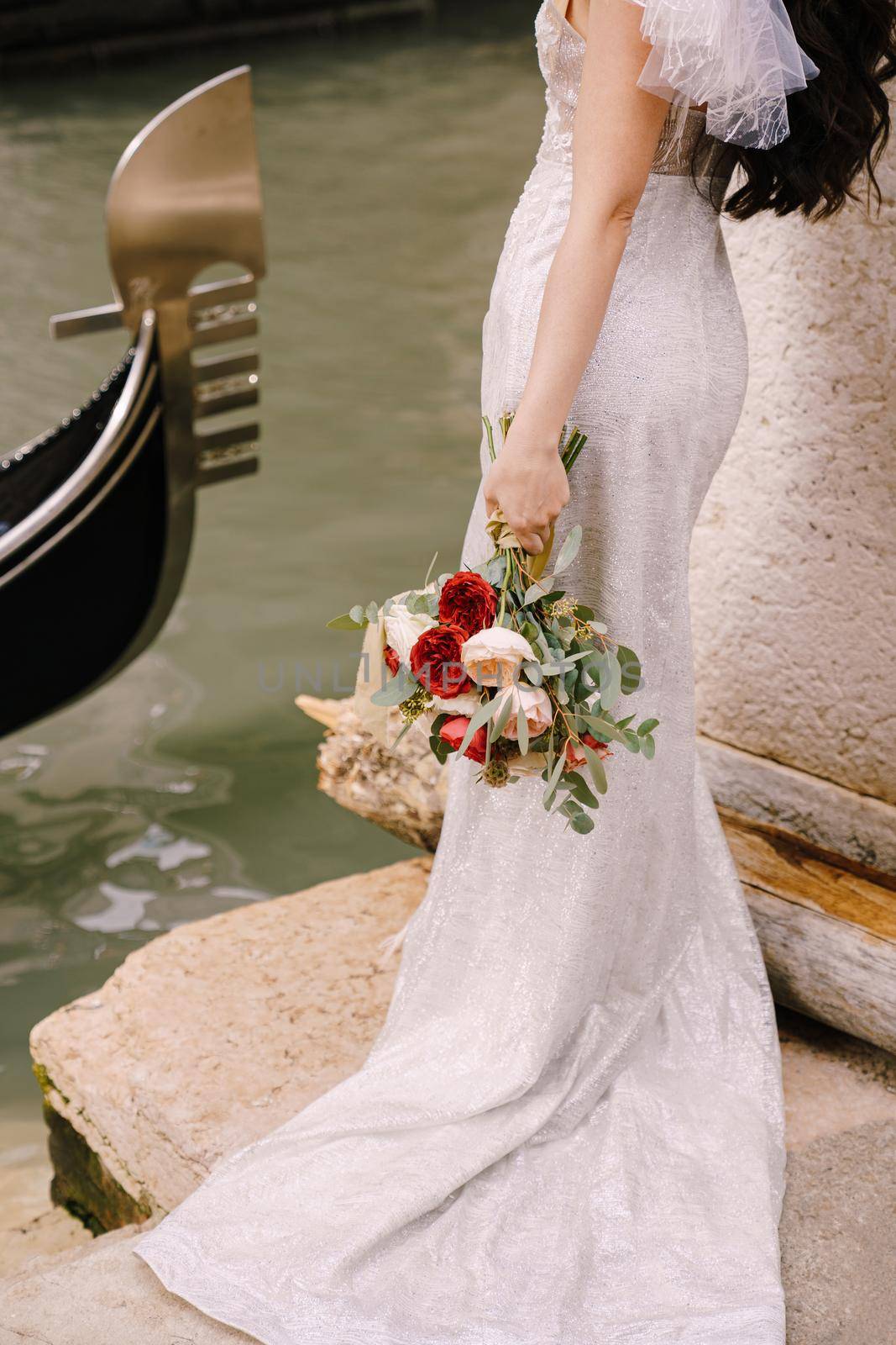 A bride in a white dress, with a train, with a bouquet of white and red roses in her hands, stands on the pier near the moored gondola in a narrow Venetian canal.
