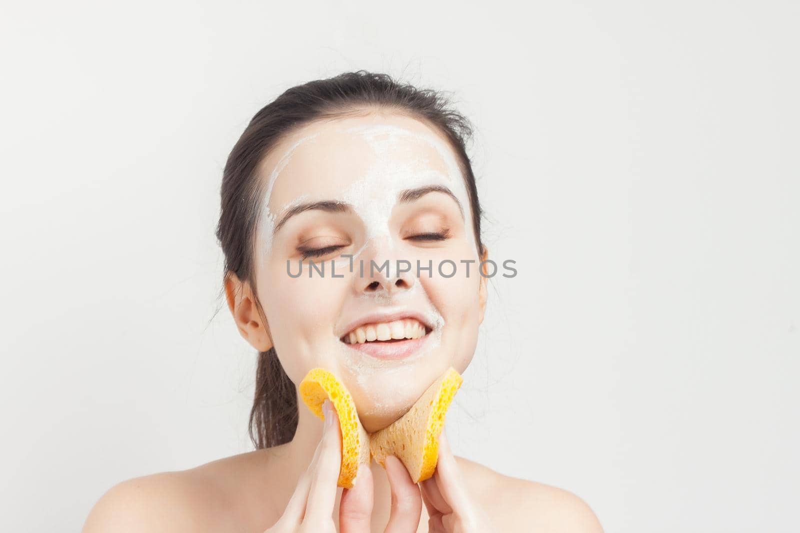 joyful woman with bare shoulders cream mask on face scrub sponges in hands. High quality photo