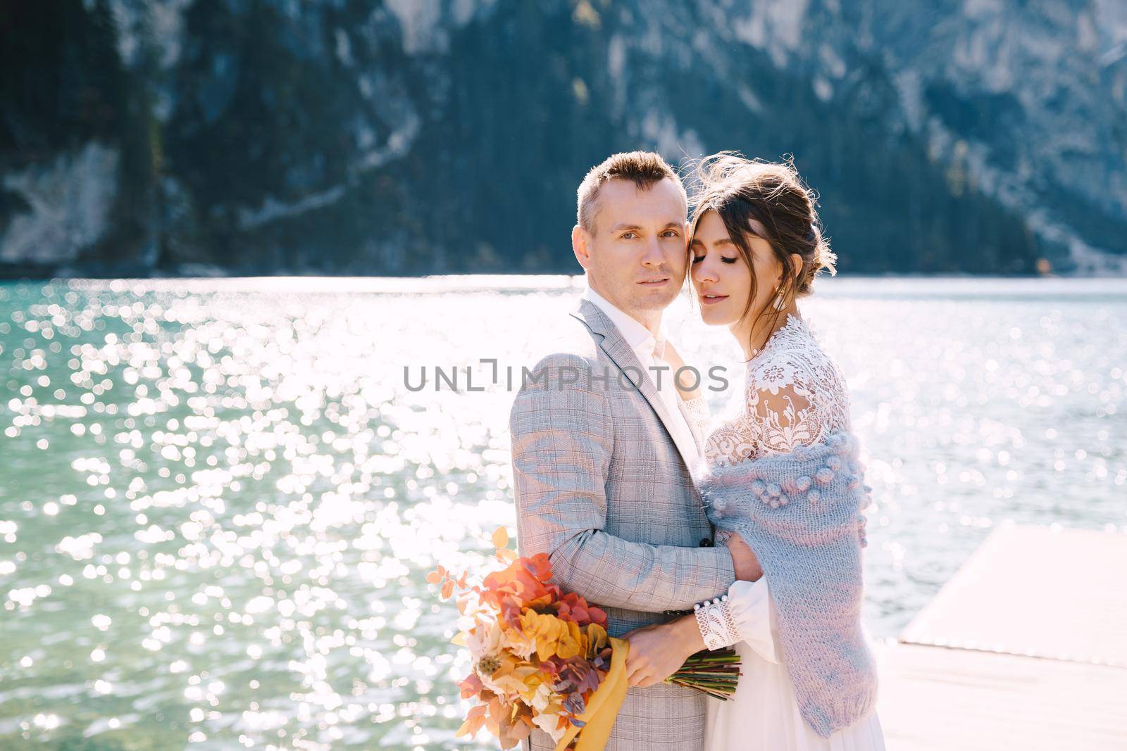 The bride and groom walk along a wooden boat dock at the Lago di Braies in Italy. Wedding in Europe, on Braies lake. Newlyweds walk, kiss, hug on a background of rocky mountains. by Nadtochiy