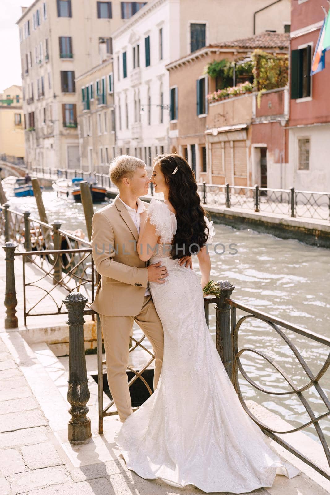 Newlyweds stand hugging on the banks of the Venice Canal. The groom hugs the bride by the waist. White wedding dress with a small beautiful train and a sandy color men's suit.