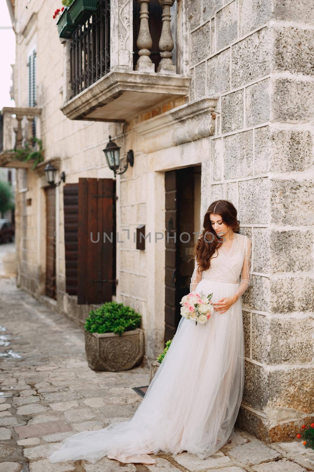 A bride with a wedding bouquet stands at a beautiful ancient building on a cozy Perast street . High quality photo