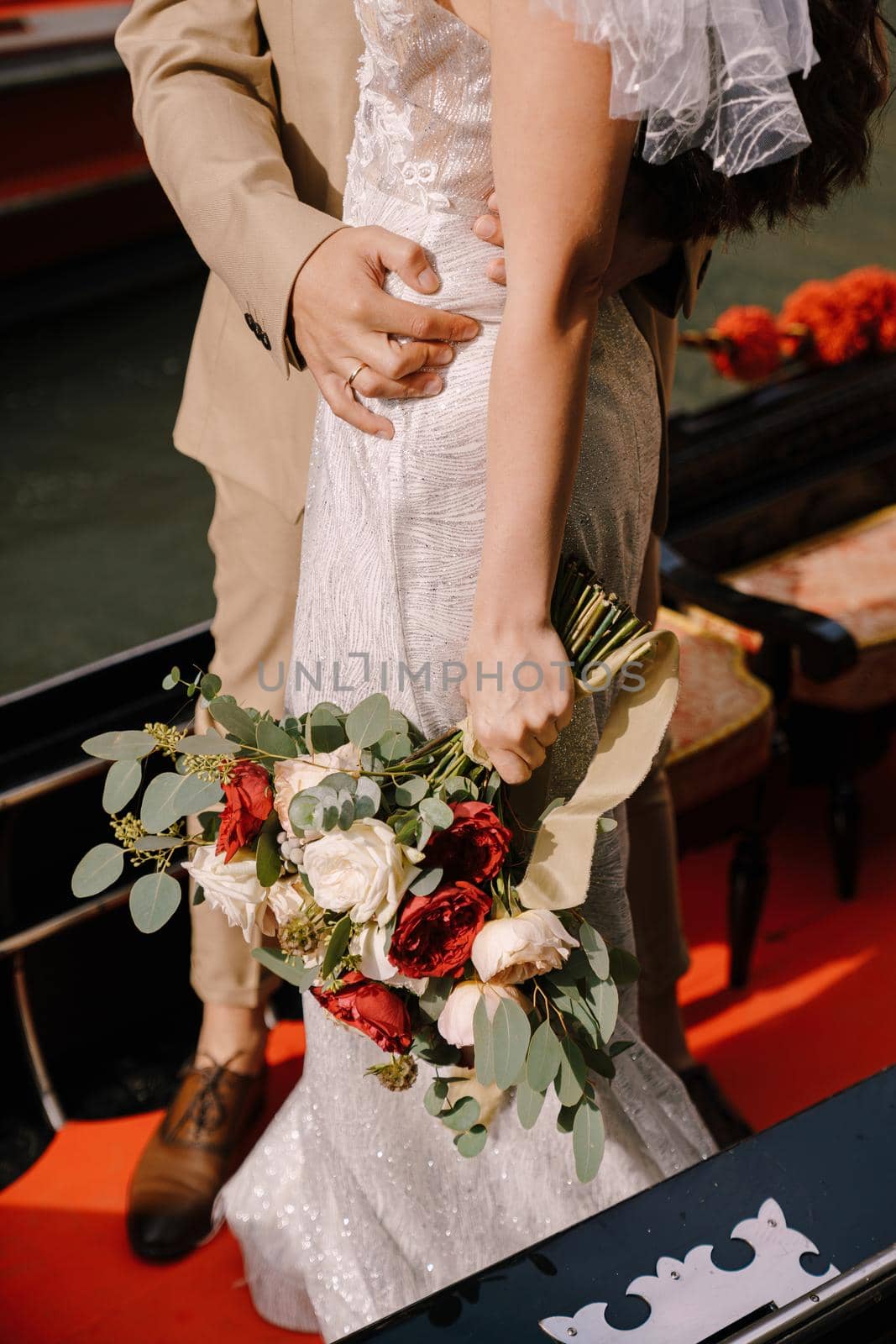 Italy wedding in Venice. Close-up - the groom hugs the bride by the waist in a classic wooden gondola. The bride holds a magnificent bouquet of white and red roses in her hand. by Nadtochiy
