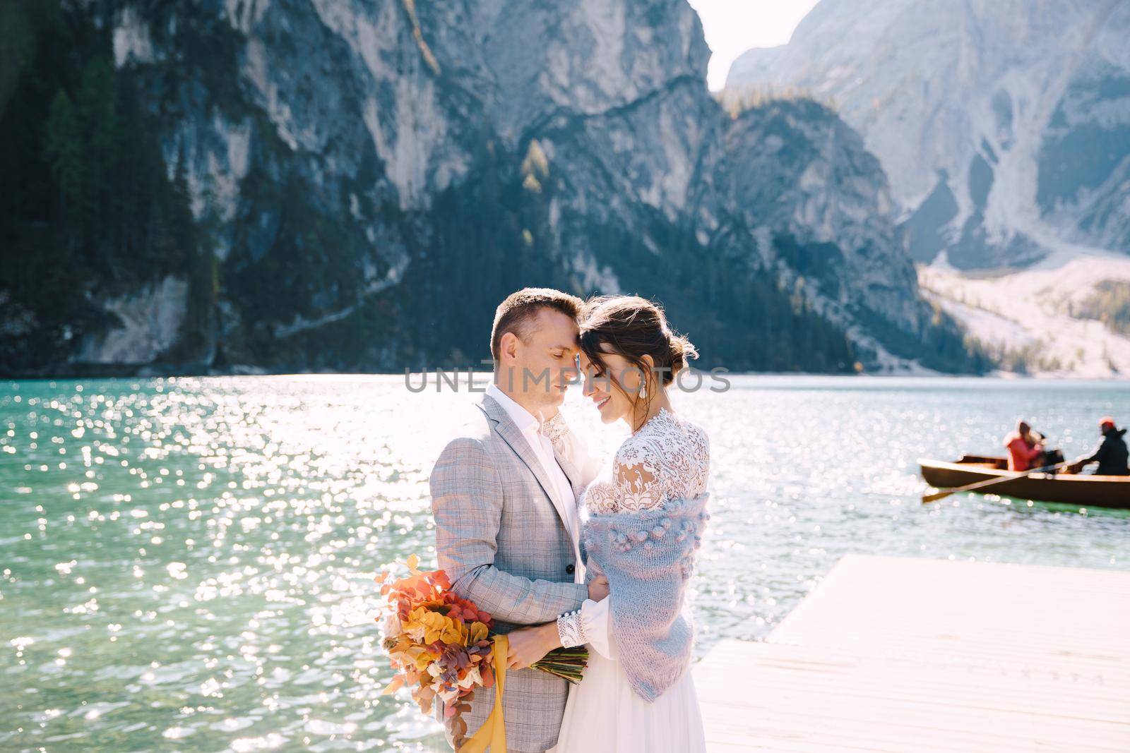The bride and groom walk on a wooden boat dock at Lago di Braies in Italy. Wedding in Europe, at Braies lake. Newlyweds walk, kiss, cuddle against the backdrop of rocky mountains.