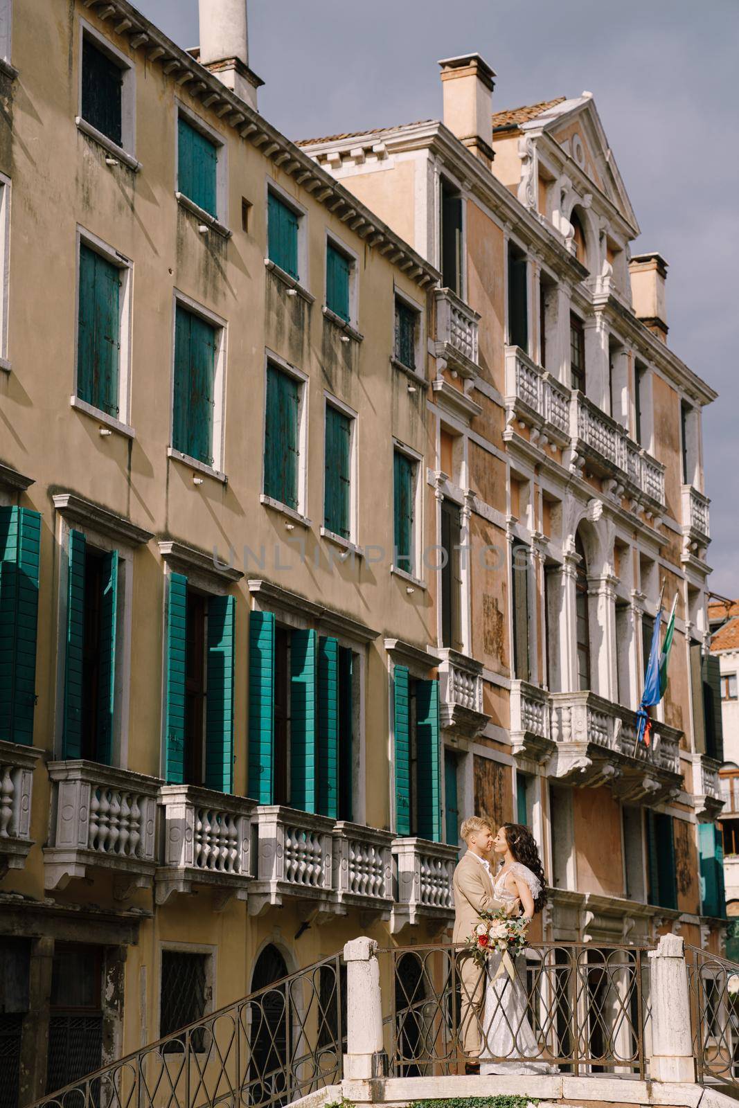 Italy wedding in Venice. The bride and groom are standing on a stone bridge over a narrow Venetian canal. Newlyweds walk around the city taking pictures on the street. by Nadtochiy