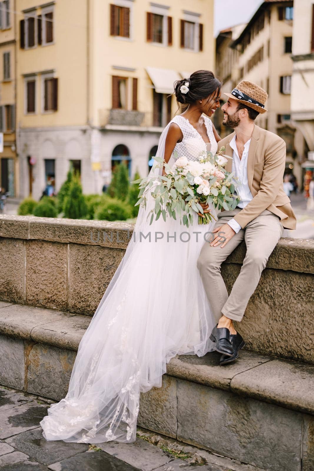 Interracial wedding couple. Wedding in Florence, Italy. African-American bride in a white dress with a long veil and a bouquet, and Caucasian groom in a sandy jacket and straw hat. by Nadtochiy