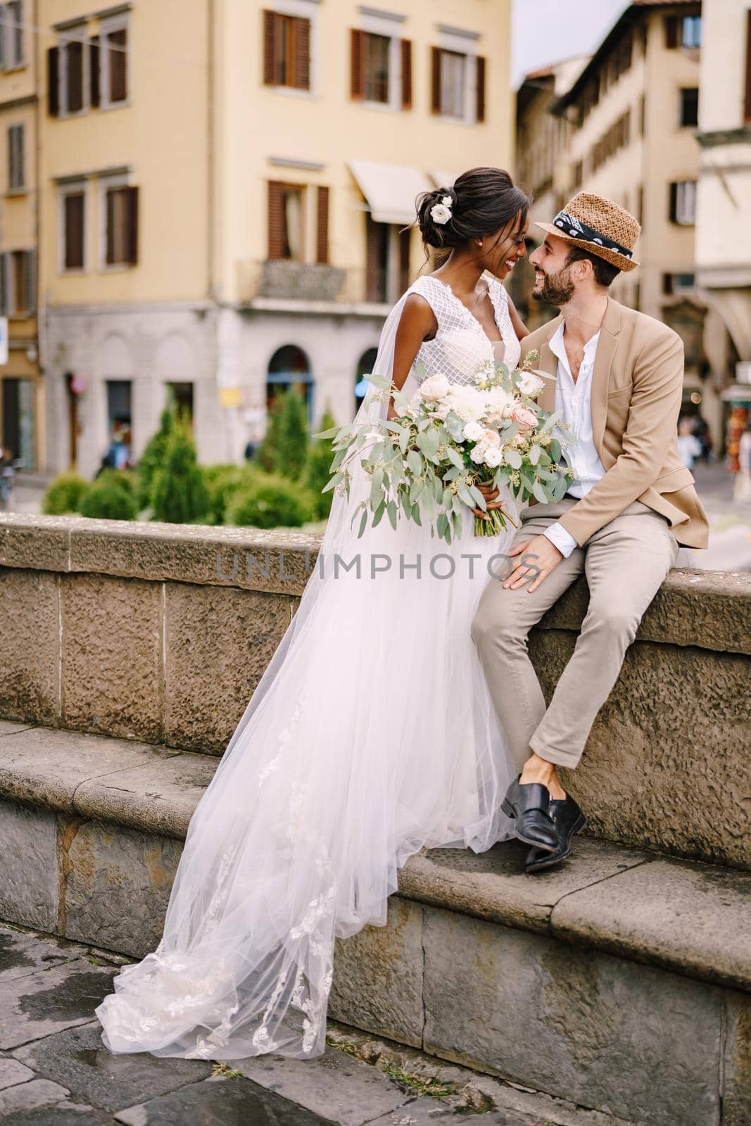 African-American bride in a white dress with a long veil and bouquet, and Caucasian groom in a sand jacket and straw hat. Interracial wedding couple. Wedding in Florence, Italy.