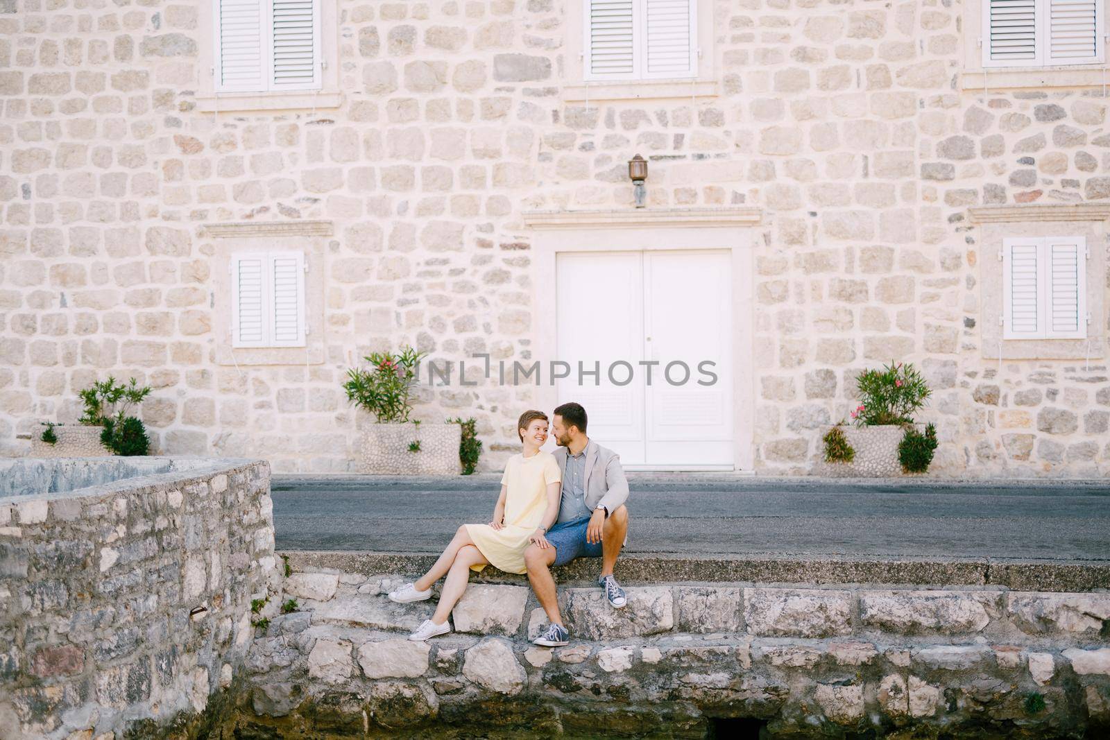 A man and a woman are sitting embracing on the road near a beautiful white house in the old town of Perast by Nadtochiy