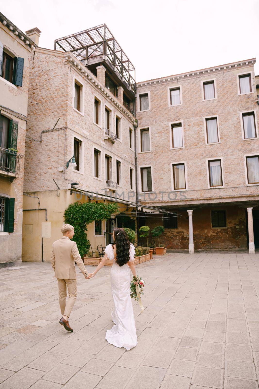 The bride and groom walk through the deserted streets of the city. The newlyweds hold hands and walk against the facade of the building.