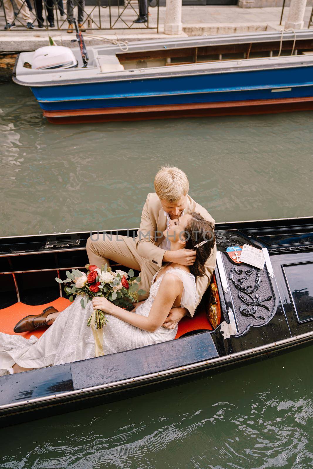 Italy wedding in Venice. The bride and groom ride in a classic wooden gondola along a narrow Venetian canal. Close-up of cuddles newlyweds. by Nadtochiy