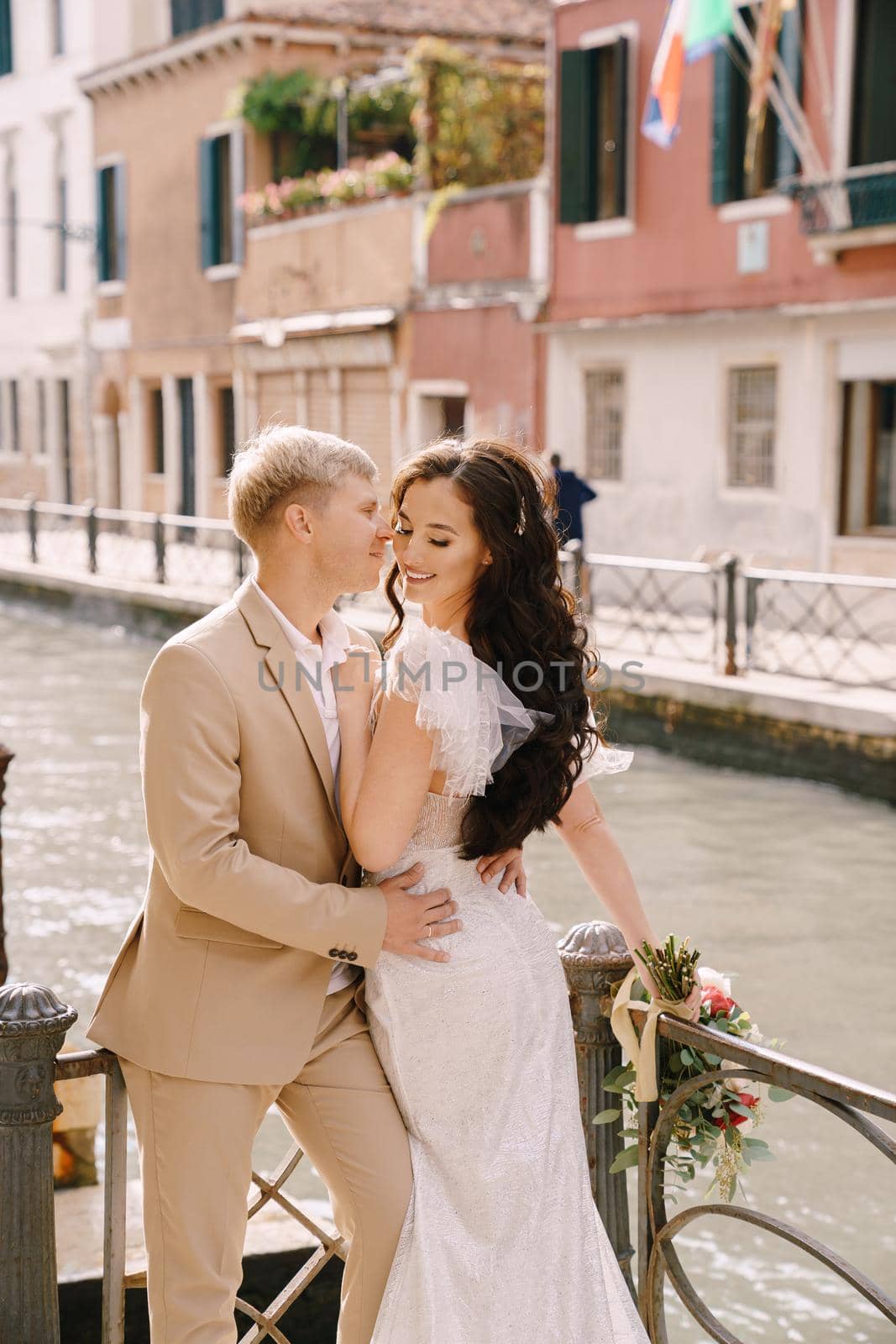 Newlyweds stand hugging on the banks of the Venice Canal. The groom hugs the bride by the waist. White wedding dress with a small beautiful train and a sandy color men's suit.