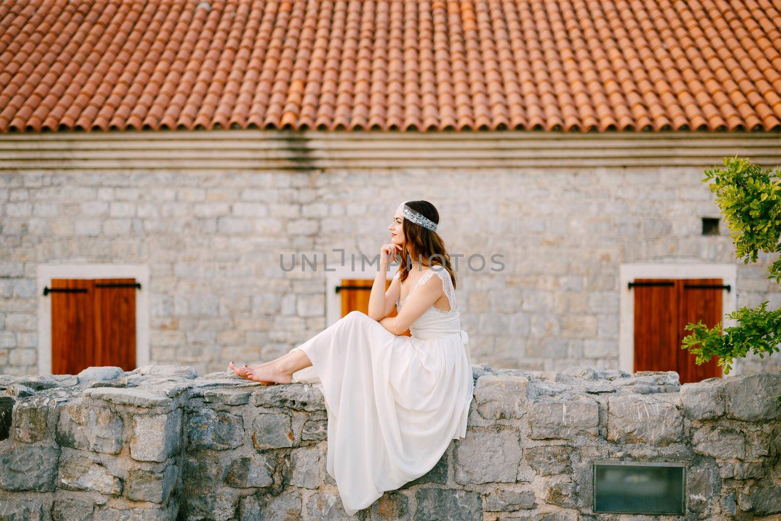 Barefoot bride sits on the stone wall of the old town of Budva . High quality photo