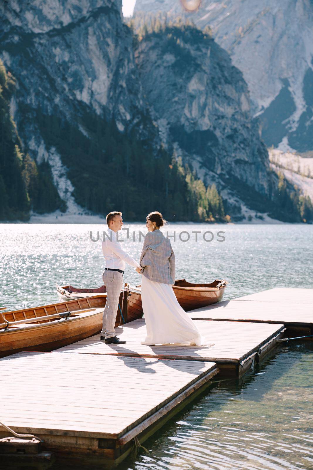 The bride and groom walk on a wooden boat dock at Lago di Braies in Italy. Wedding in Europe, at Braies lake. Newlyweds walk, kiss, cuddle against the backdrop of rocky mountains.