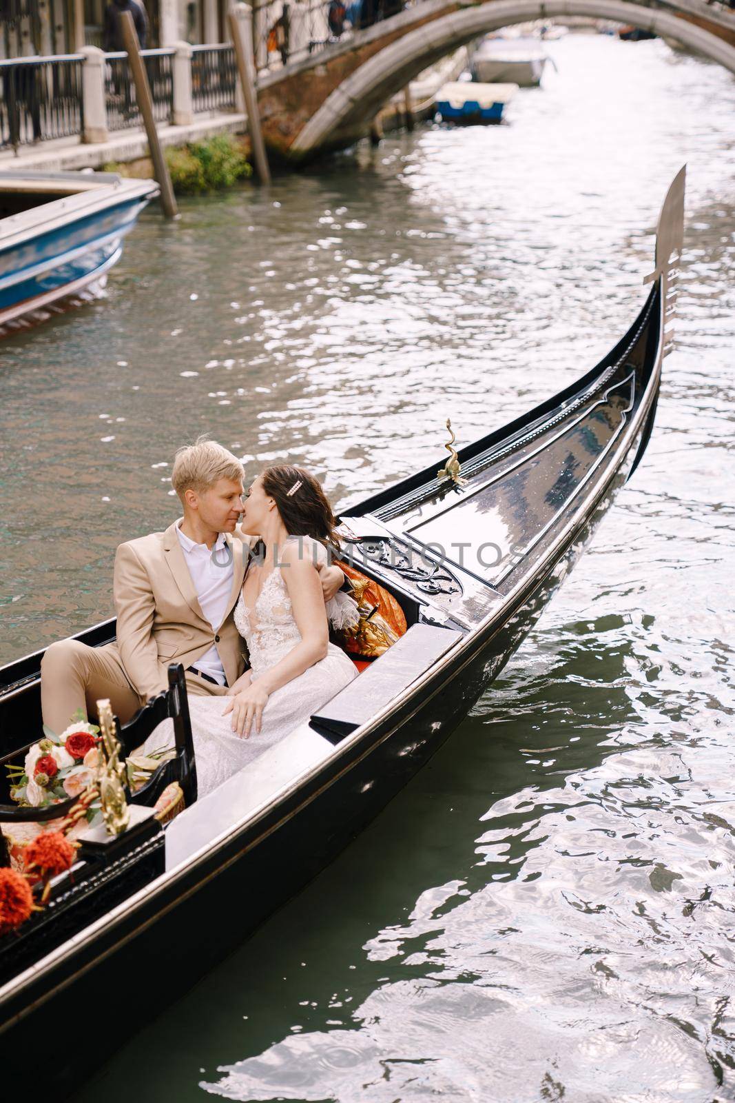 Italy wedding in Venice. The bride and groom ride in a classic wooden gondola along a narrow Venetian canal. Close-up of cuddles newlyweds. by Nadtochiy