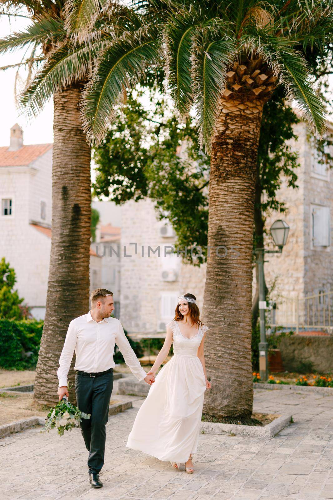 The bride and groom walk hand in hand through the old town of Budva, next to beautiful tall palm trees by Nadtochiy