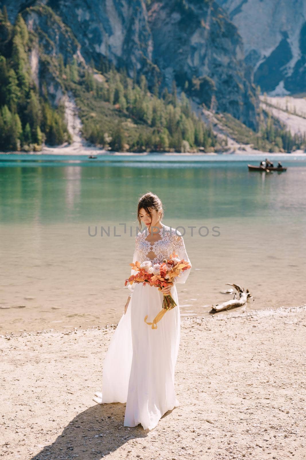 Beautiful bride in a white dress with sleeves and lace, with a yellow autumn bouquet of dried flowers and peony roses, on the Lago di Braies in Italy. Destination wedding in Europe, on Braies lake. by Nadtochiy