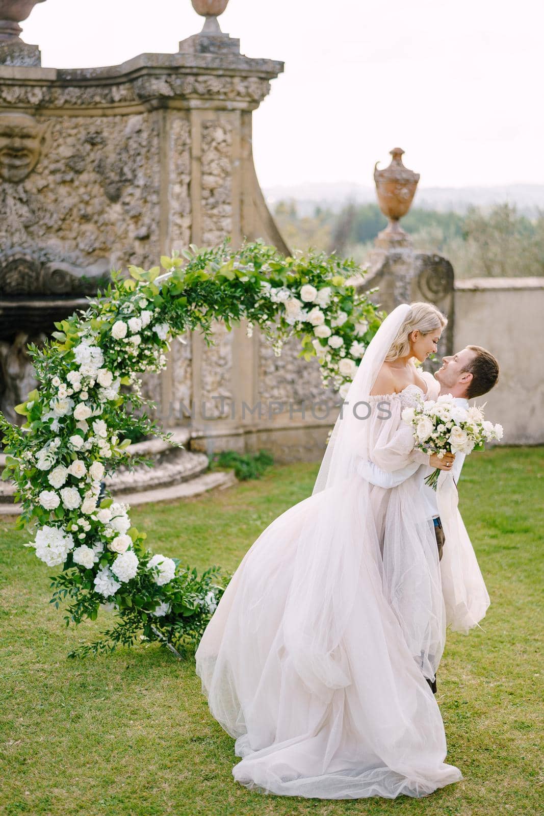 The groom is circling the bride in her arms. Round wedding arch decorated with white flowers and greenery in front of an ancient Italian architecture. Wedding at an old winery villa in Tuscany, Italy.