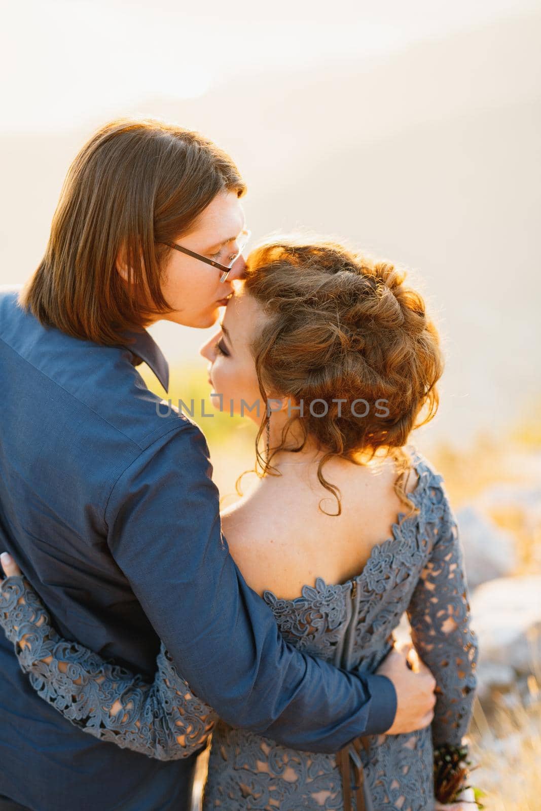 The bride and groom embracing on the Lovcen mountain, the groom kisses the bride on the forehead, back view by Nadtochiy