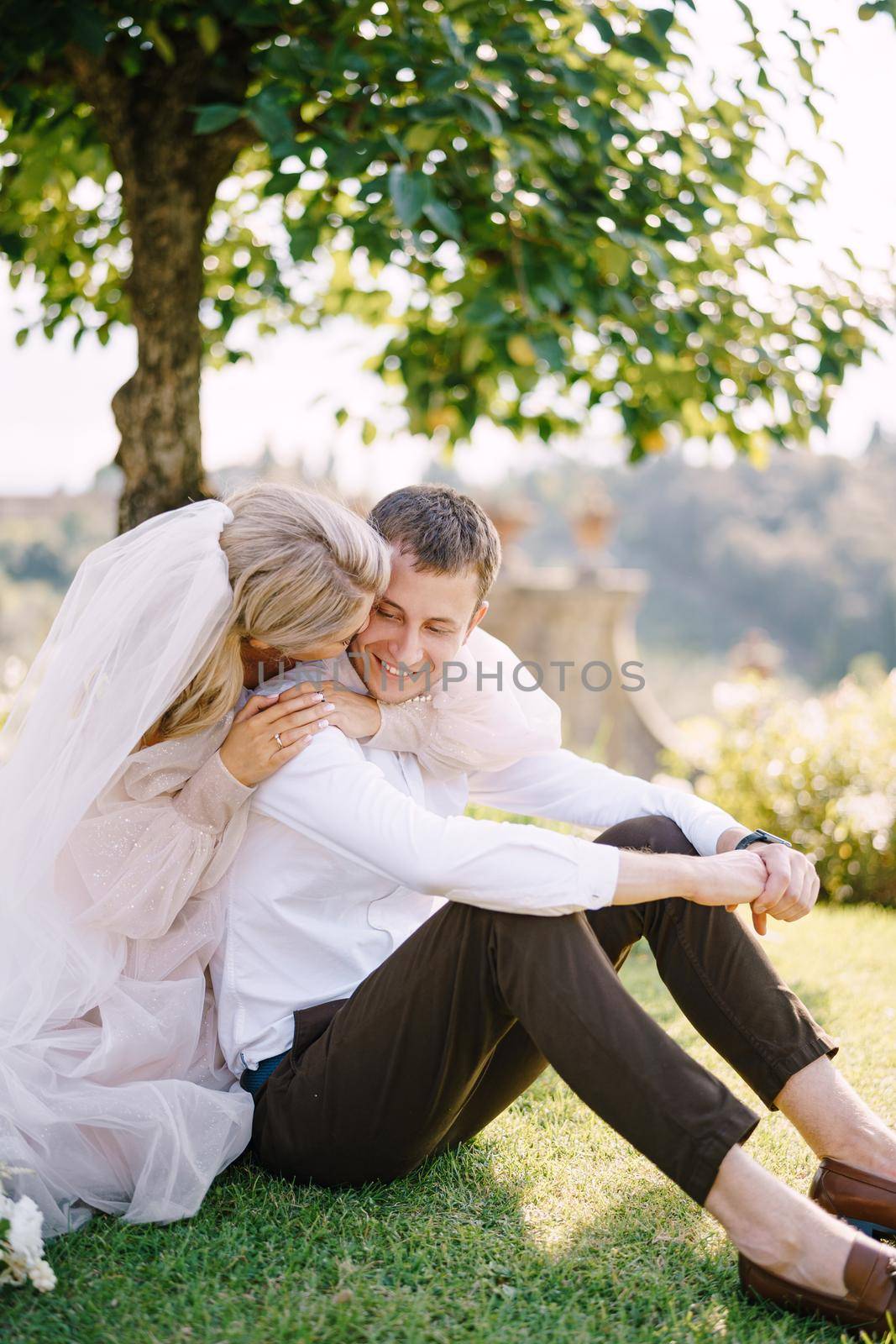 Wedding in Florence, Italy, in an old villa-winery. A wedding couple is sitting on the grass in the garden under a tree, the bride is hugging the groom. by Nadtochiy