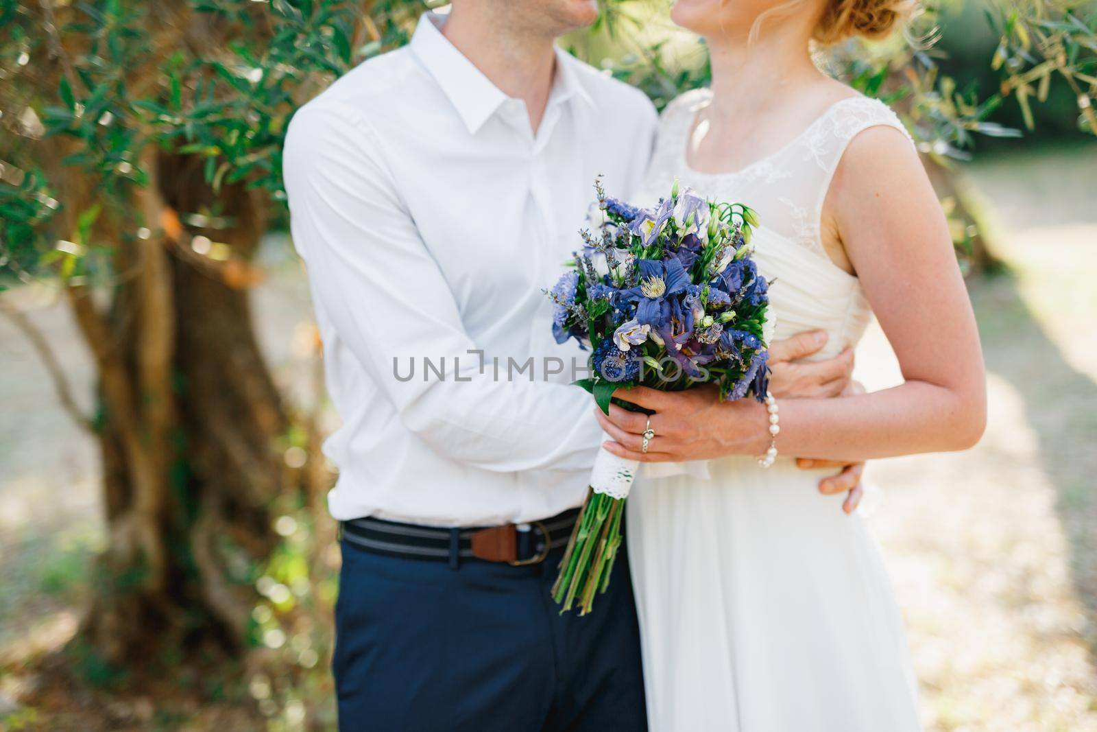 Bride with a bouquet of blue flowers and the groom embracing tenderly in the olive grove by Nadtochiy