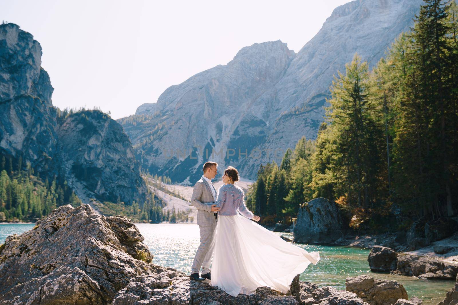 The bride and groom are standing on stones overlooking the Lago di Braies in Italy. Destination wedding in Europe, on Braies lake. Loving newlyweds walk against the backdrop of amazing nature. by Nadtochiy