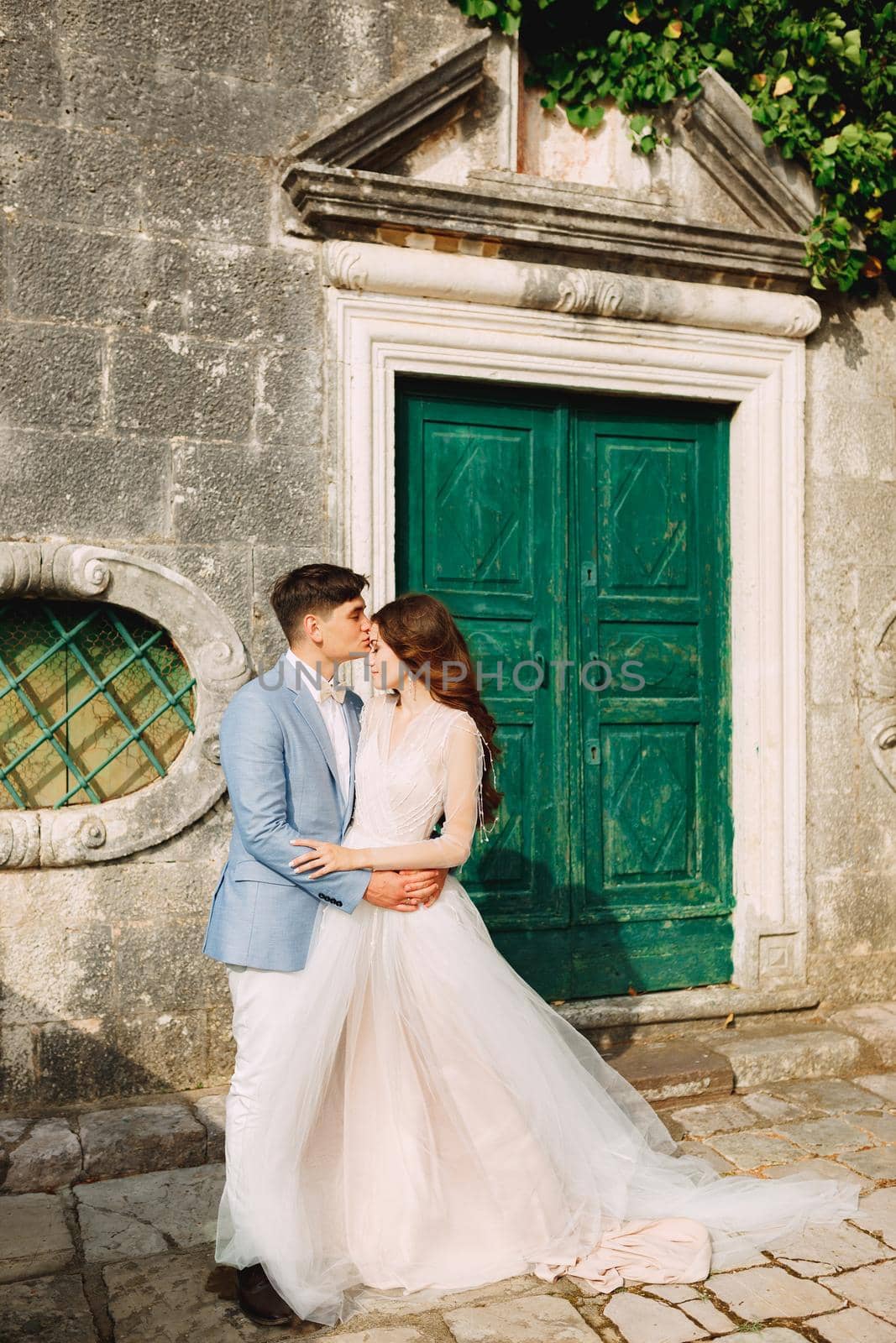 The bride and groom are embracing at the wooden door of an ancient building in Perast, the groom kisses the bride on the forehead by Nadtochiy
