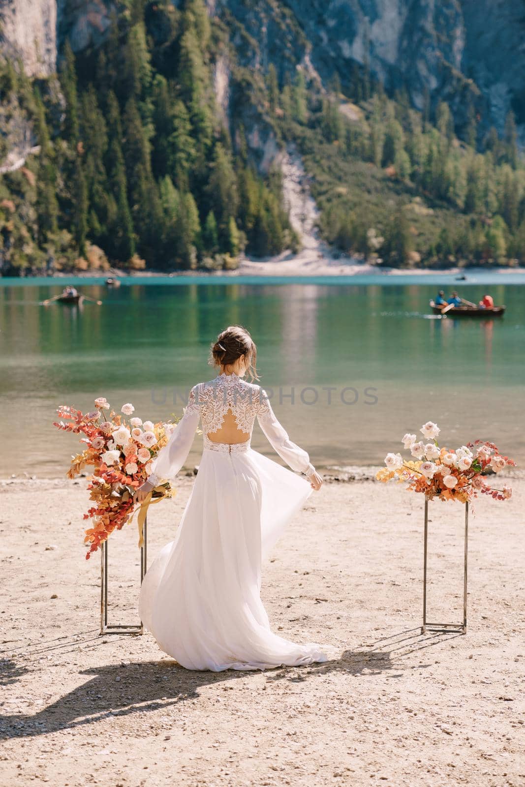 Beautiful bride in a white dress with sleeves and lace, with a yellow autumn bouquet against backdrop of arch for ceremony, at Lago di Braies in Italy. Destination wedding in Europe, at Braies lake.