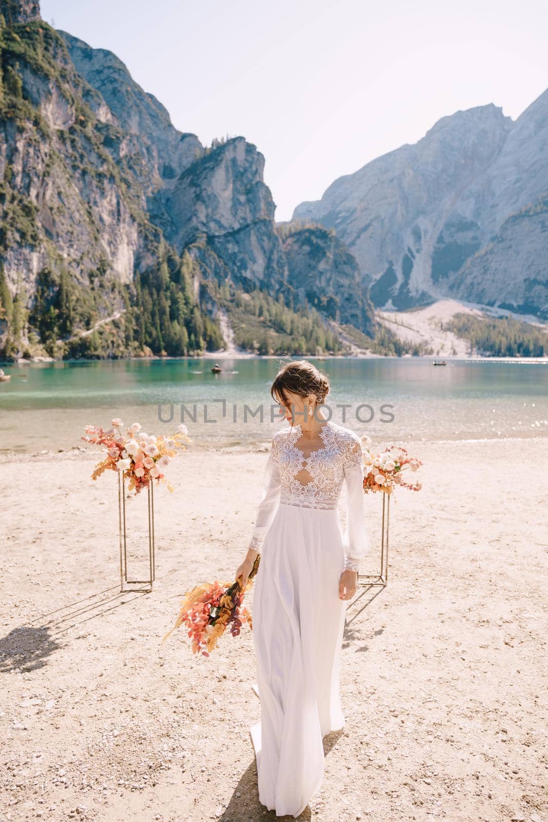 Beautiful bride in a white dress with sleeves and lace, with a yellow autumn bouquet on background of the arch for ceremony, at Lago di Braies in Italy. Destination wedding in Europe, on Braies lake. by Nadtochiy