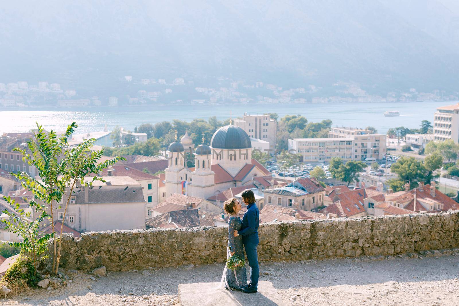 The bride and groom hug on the observation deck with a picturesque view of the old town of Kotor and the Bay of Kotor by Nadtochiy