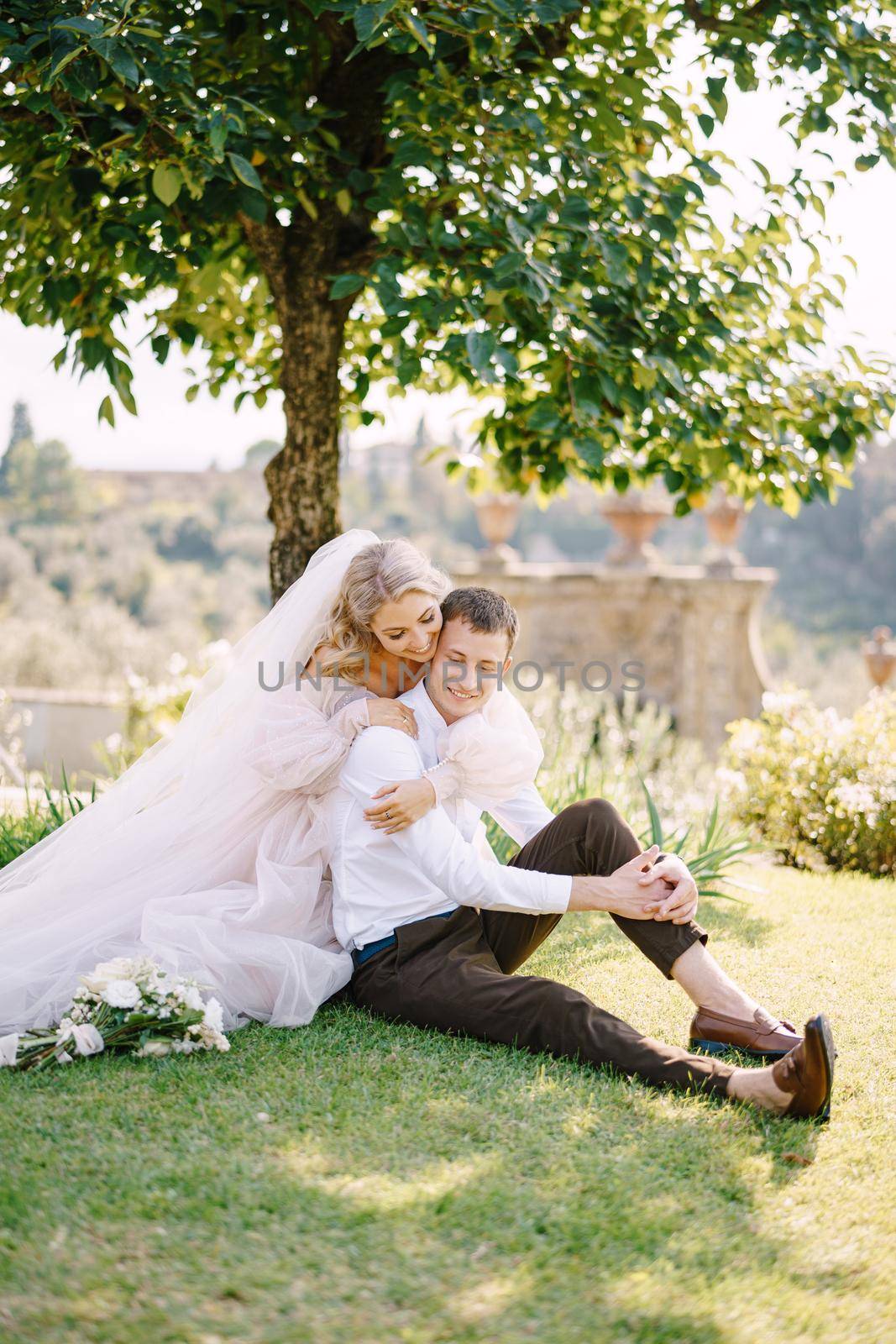 Wedding in Florence, Italy, in an old villa-winery. A wedding couple is sitting on the grass in the garden under a tree, the bride is hugging the groom. by Nadtochiy
