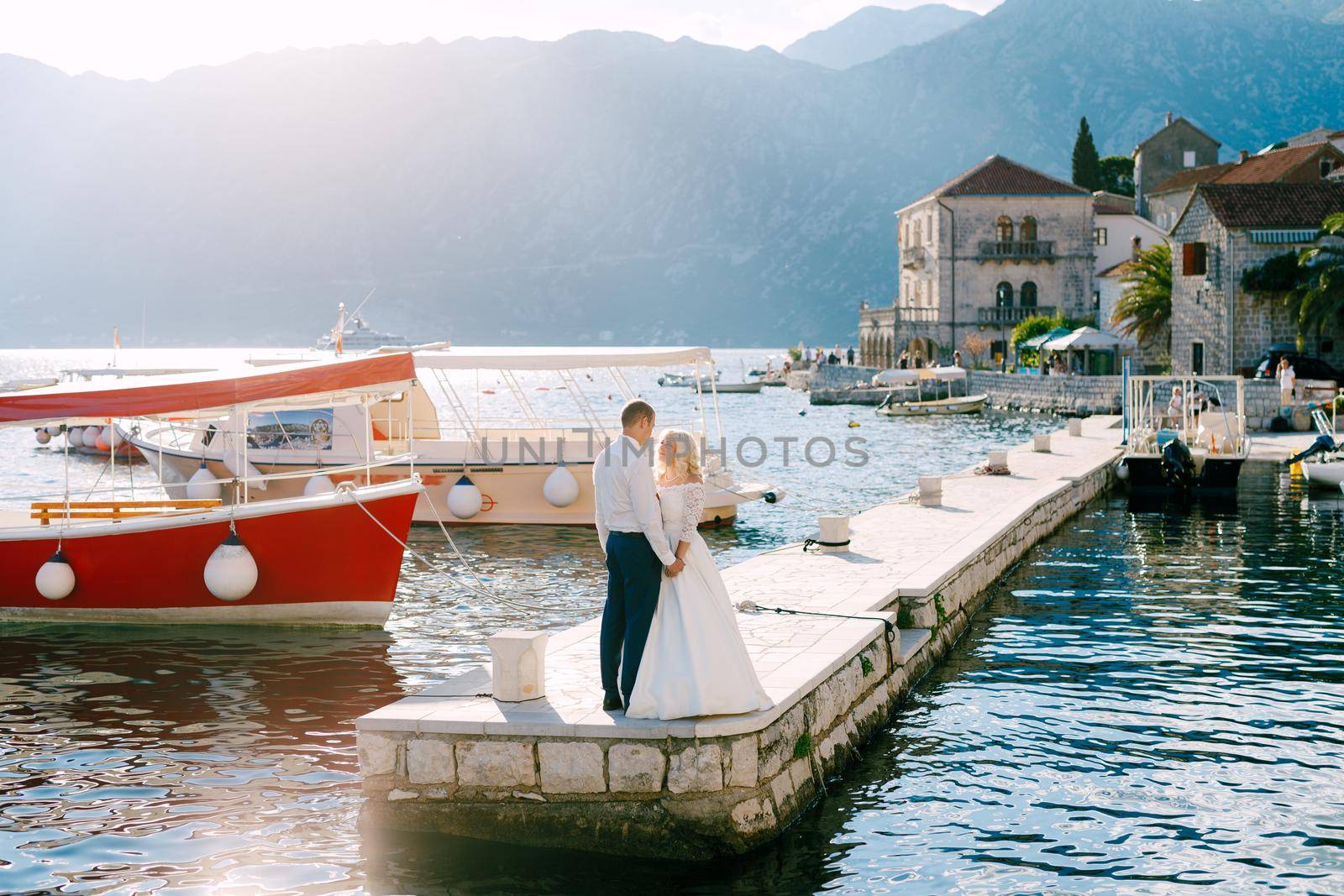 The bride and groom stand on the pier near Perast, holding hands and looking into each other's eyes, next to them boats . High quality photo