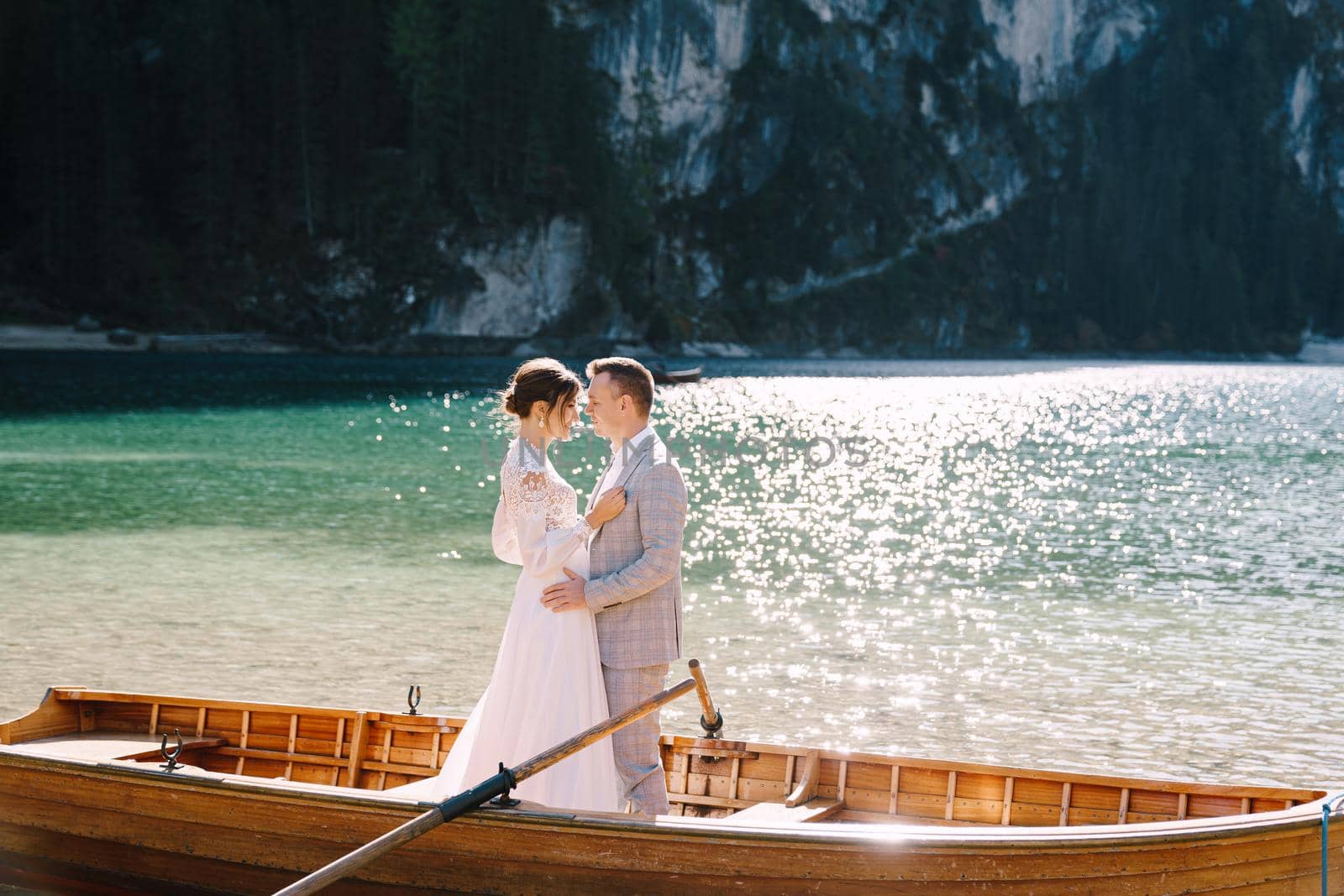 Bride and groom sailing in wooden boat, with oars at Lago di Braies lake in Italy. Wedding in Europe - Newlyweds are standing embracing in a boat. by Nadtochiy
