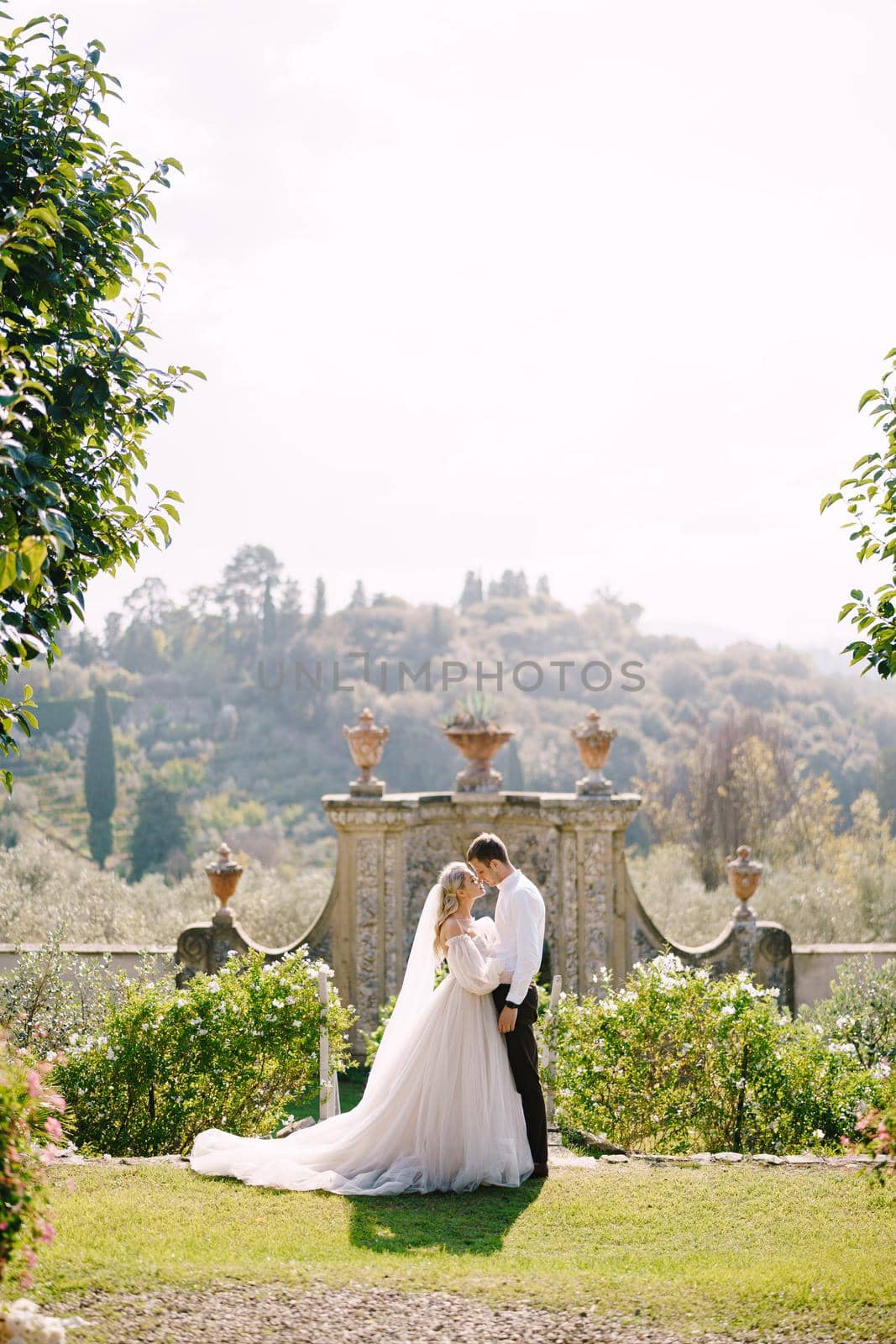 The bride and groom walk in the park. Wedding at an old winery villa in Tuscany, Italy. Round wedding arch decorated with white flowers and greenery in front of an ancient Italian architecture. by Nadtochiy