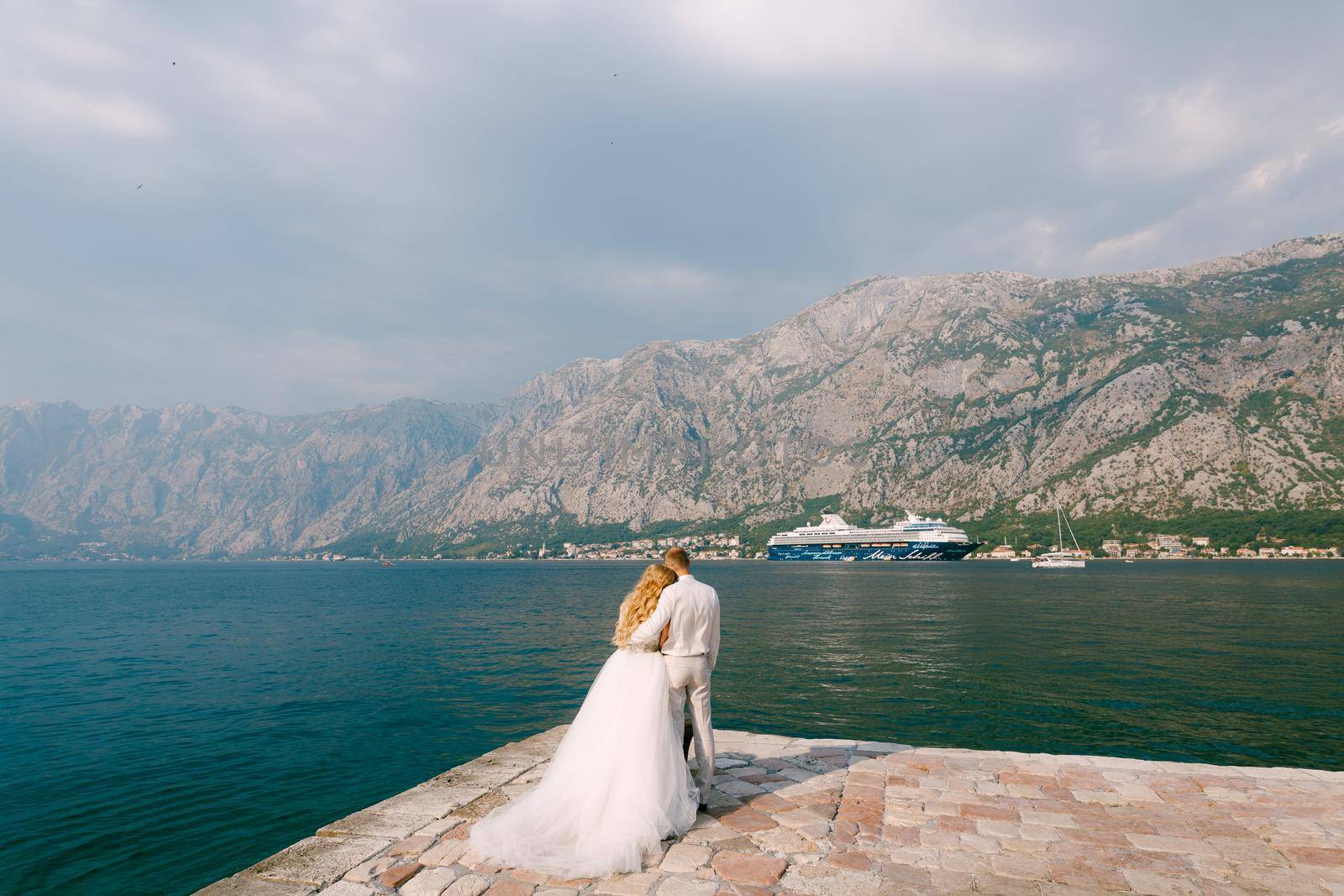 Prcanj, Montenegro - 03 june 2019: The bride and groom are embracing on the pier in the Bay of Kotor, a cruise ship is sailing in front of them, back view by Nadtochiy